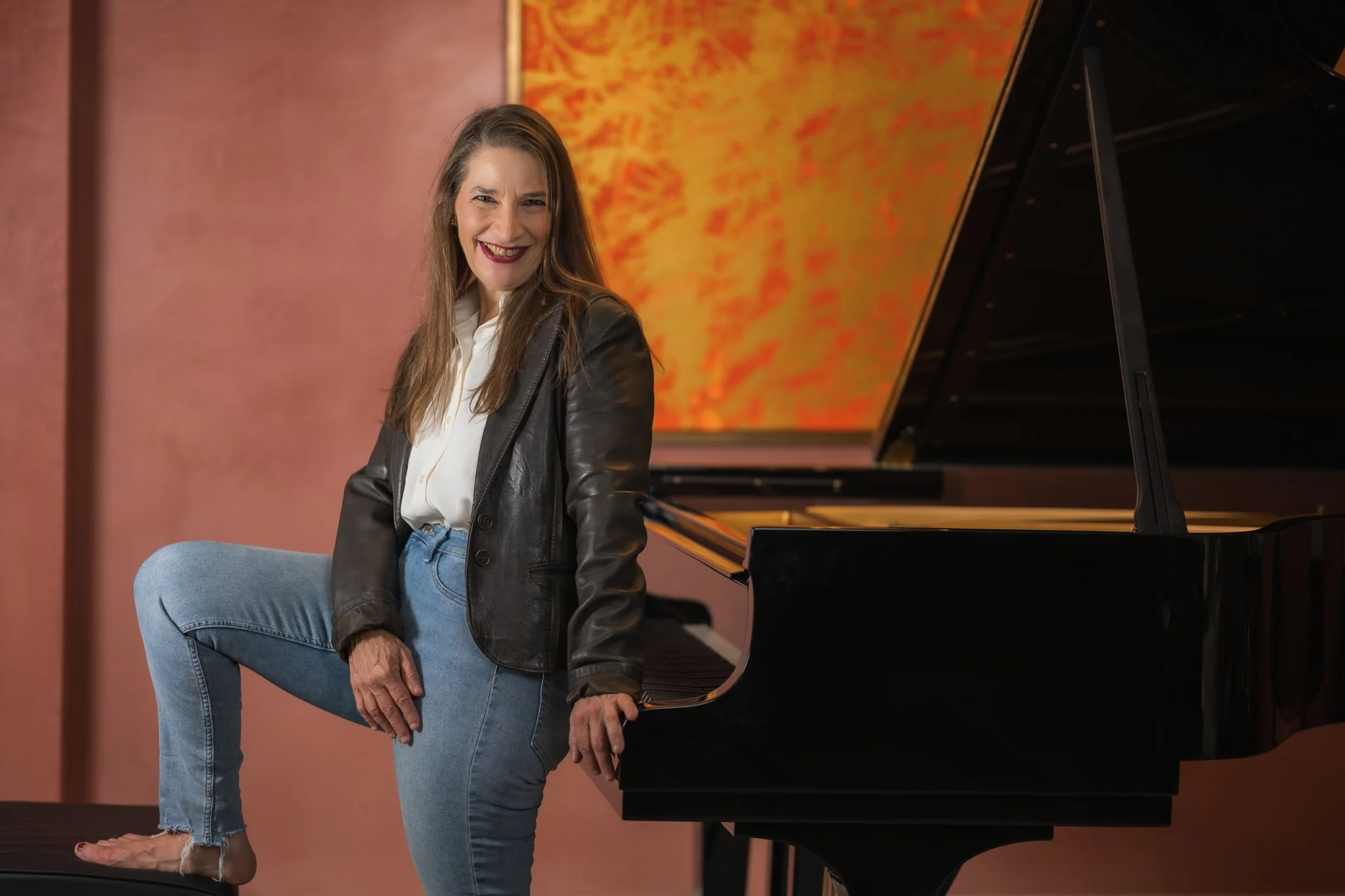 Nadia Weintraub assise près d'un piano à queue dans un espace intérieur, souriante, portant une veste en cuir et un pantalon en jean.