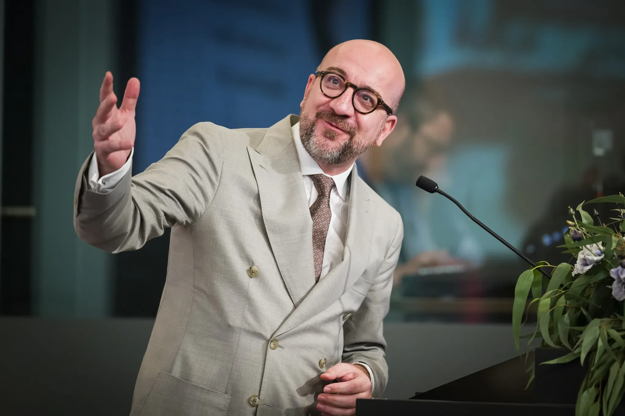 Un intervenant en veste claire et lunettes s’exprime au microphone lors d’une conférence, une main levée.