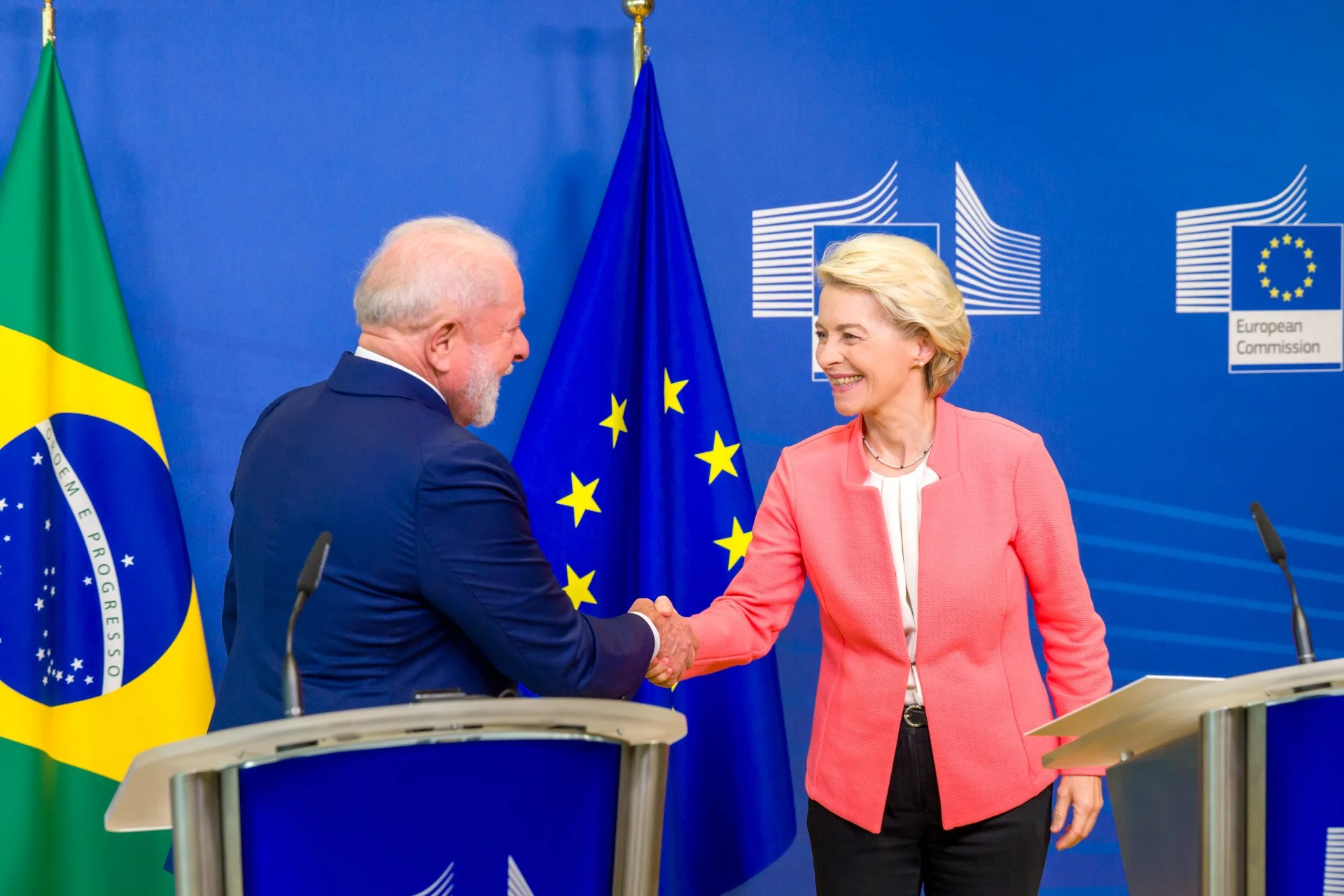 Une femme blonde habillée en rose et un homme plus âgé avec une barbe, habillé en costume foncé, souriant en se serrant la main lors d'une rencontre officielle avec les drapeaux de l'Union européenne, du Brésil et l'emblème de la Commission européenn