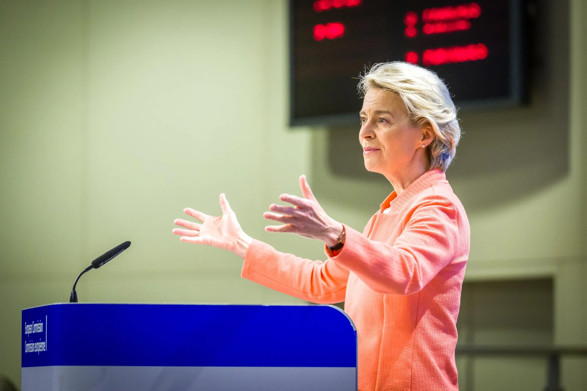 Photo professionnelle de Ursula von der Leyen prise pendant un événement institutionnel à la Commission européenne à Bruxxels. On la voit ici de 3/4 profil, écartant les bras devant un lutriin et en tailleur rose.