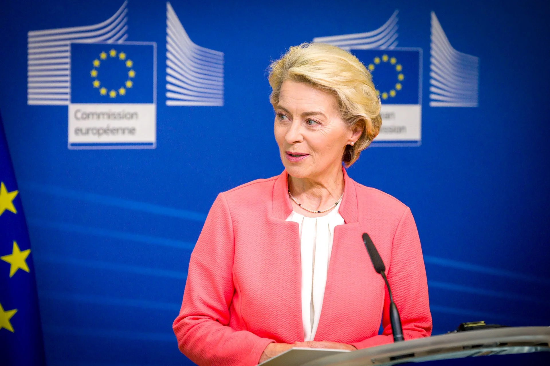 Une femme politique blonde en train de parler lors d'une conférence, avec un drapeau européen en arrière-plan