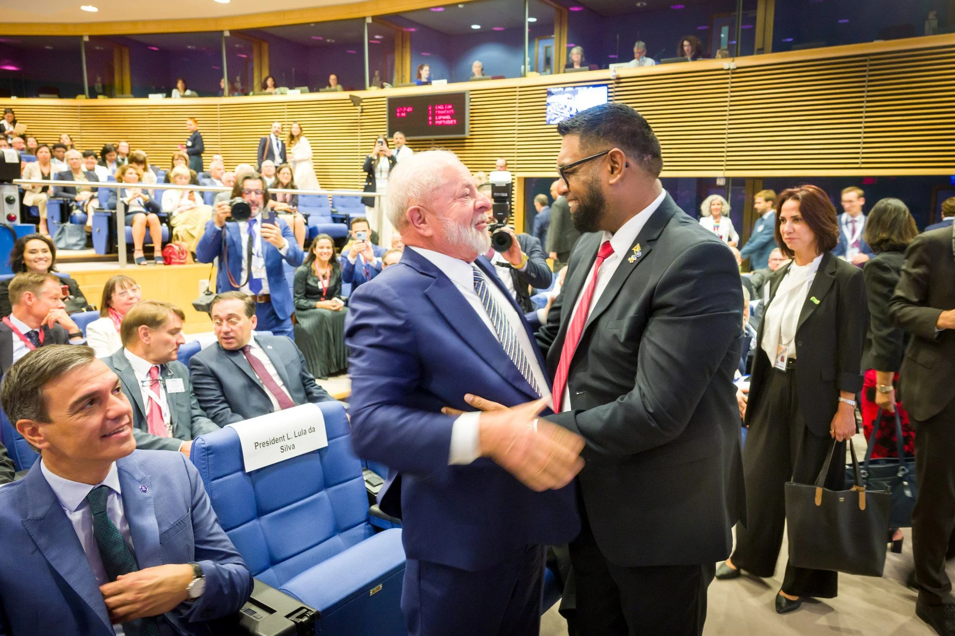 Deux hommes en costume se saluant et souriant dans une salle de conférence remplie de personnes, plusieurs photographes prennent des photos.