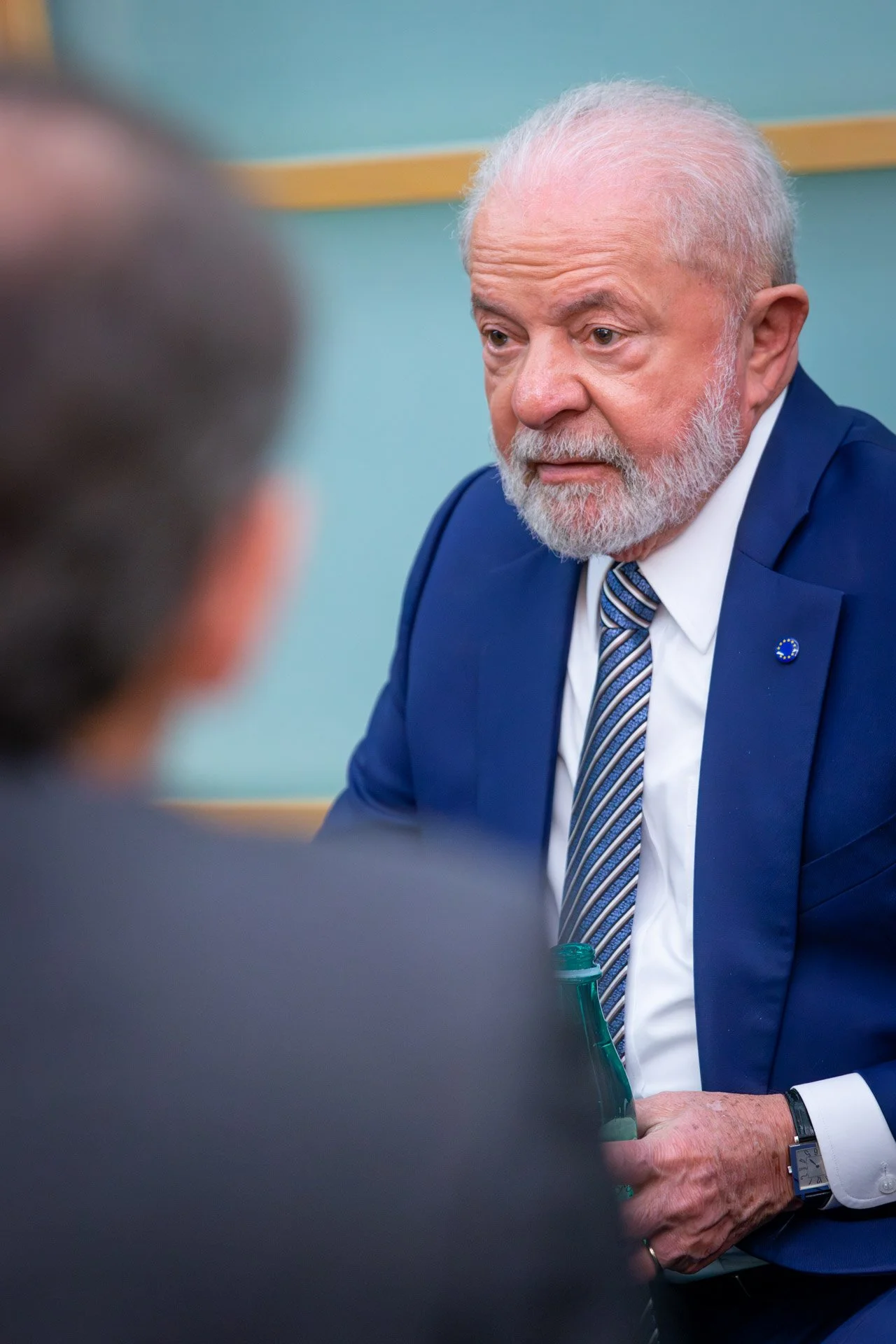 Un homme barbu en costume bleu avec une cravate à rayures, assis et tenant une bouteille verte, lors d'une discussion formelle.