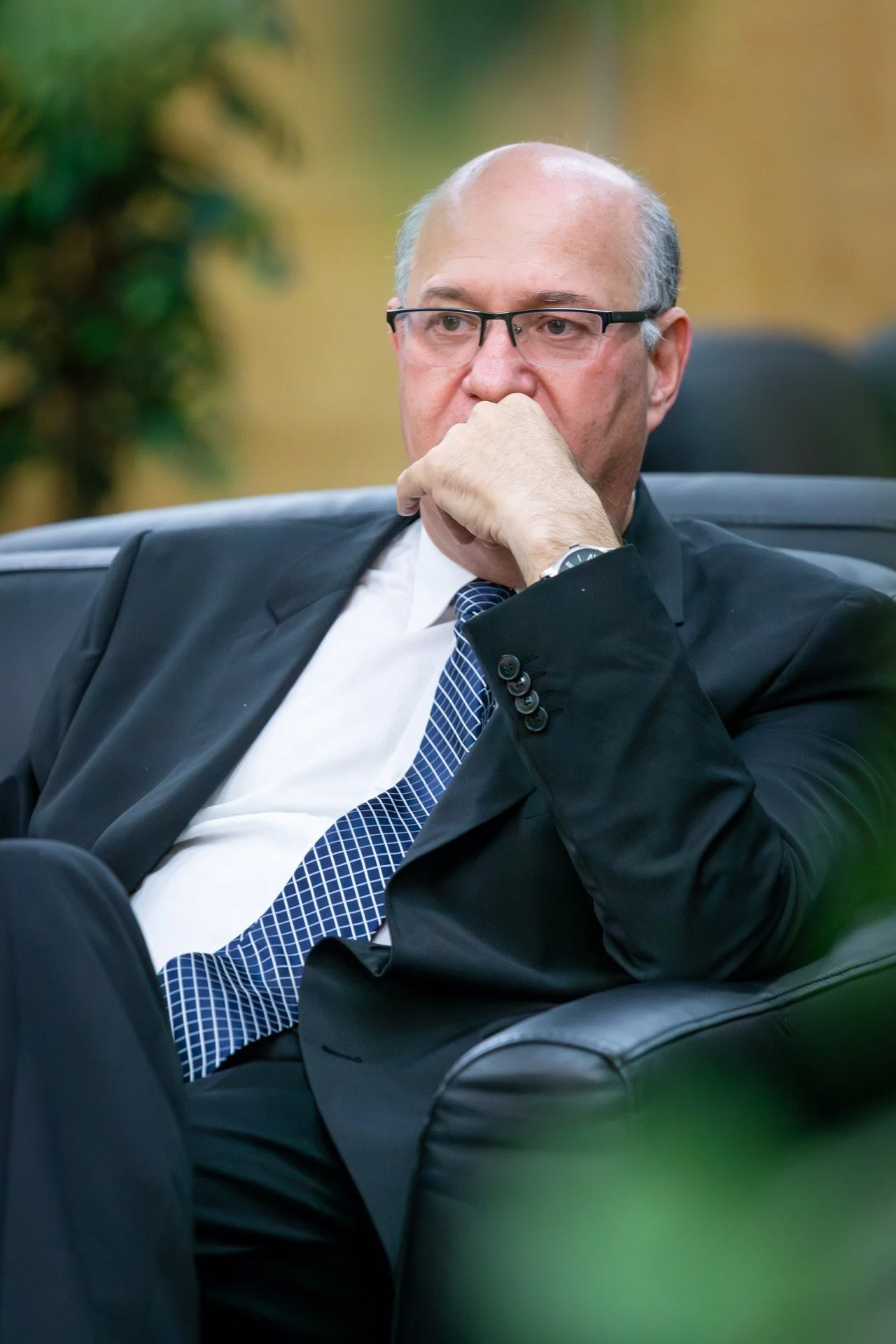 Homme en costume noir assis pensif, portant des lunettes et une cravate bleue.