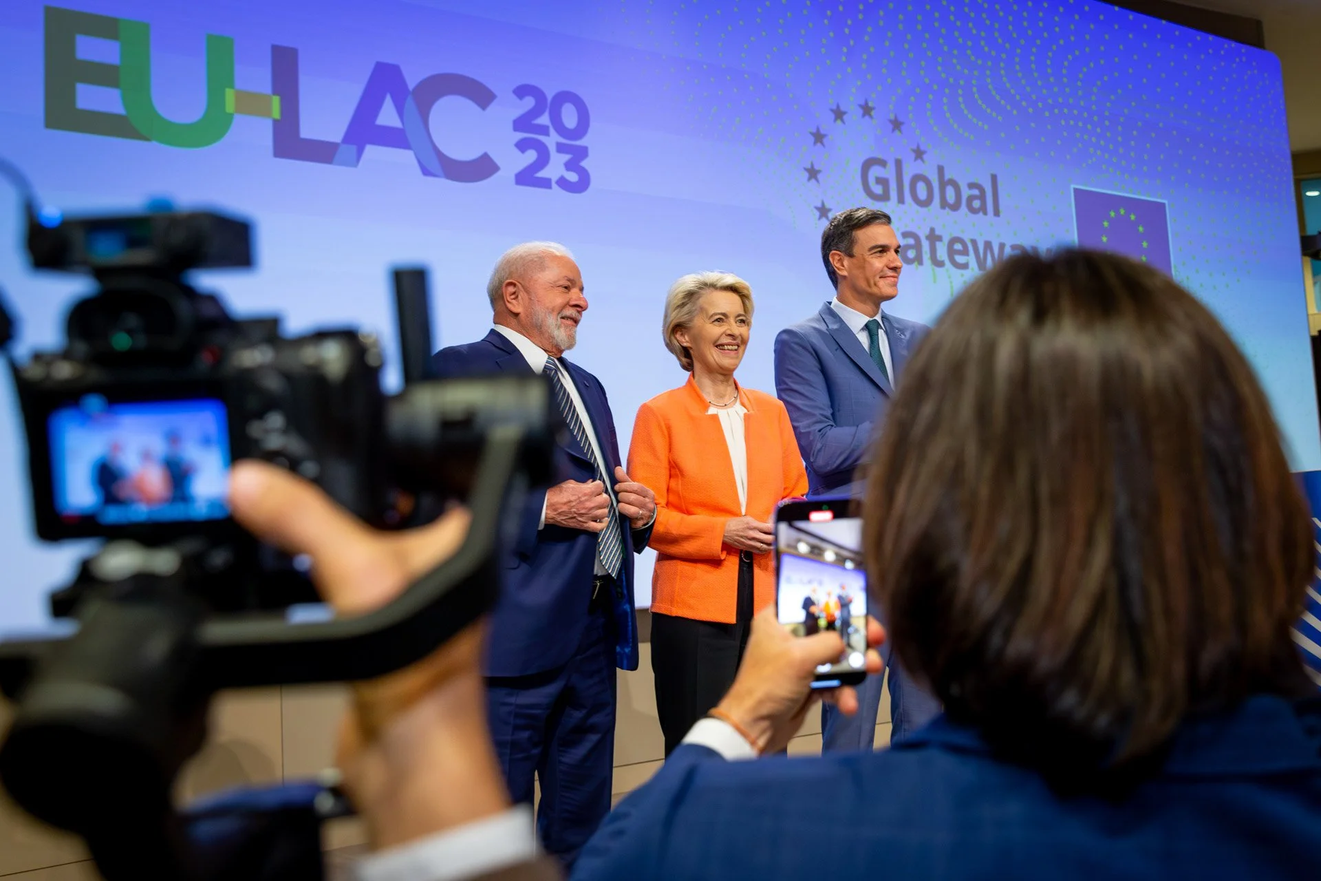 Trois personnes sont debout devant une affiche avec "EU-LAC 2023" et "Global Gateway", lors d'un événement officiel, capturées par des caméras.