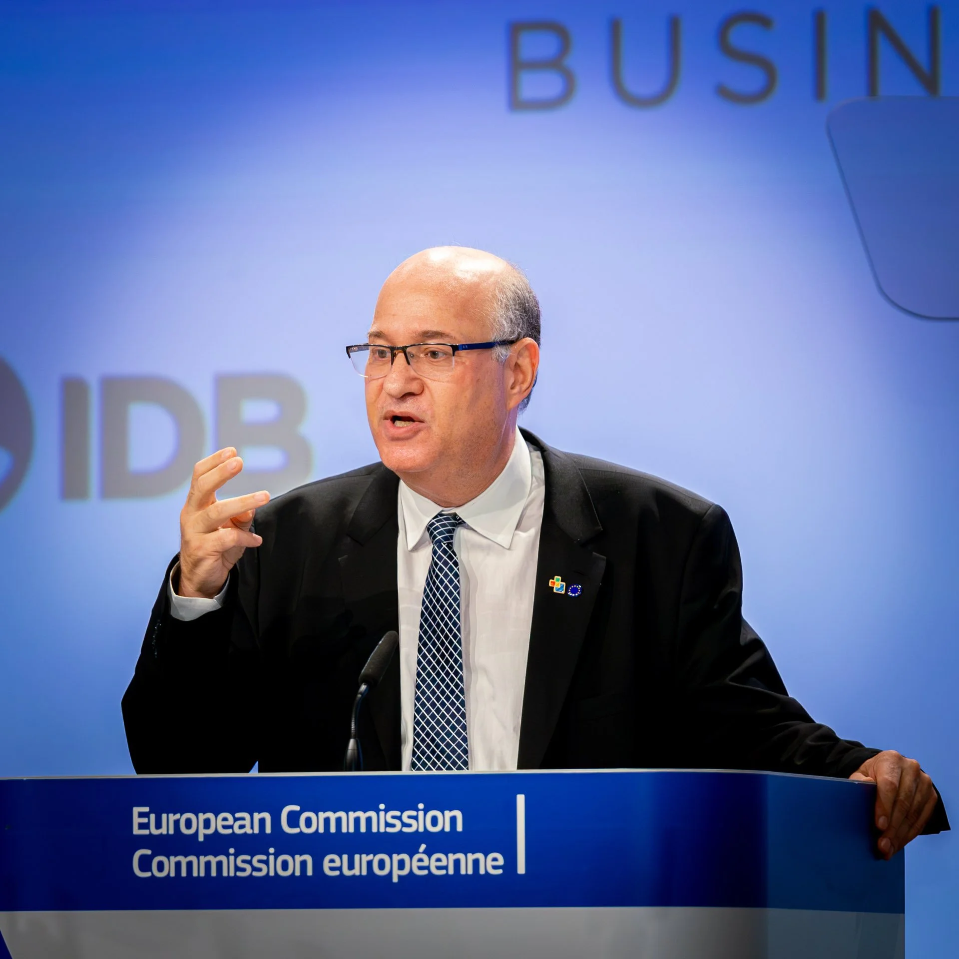 Un homme en costume s'exprimant derrière un pupitre de la Commission européenne, avec un micro et un fond bleu avec du texte partiellement visible.