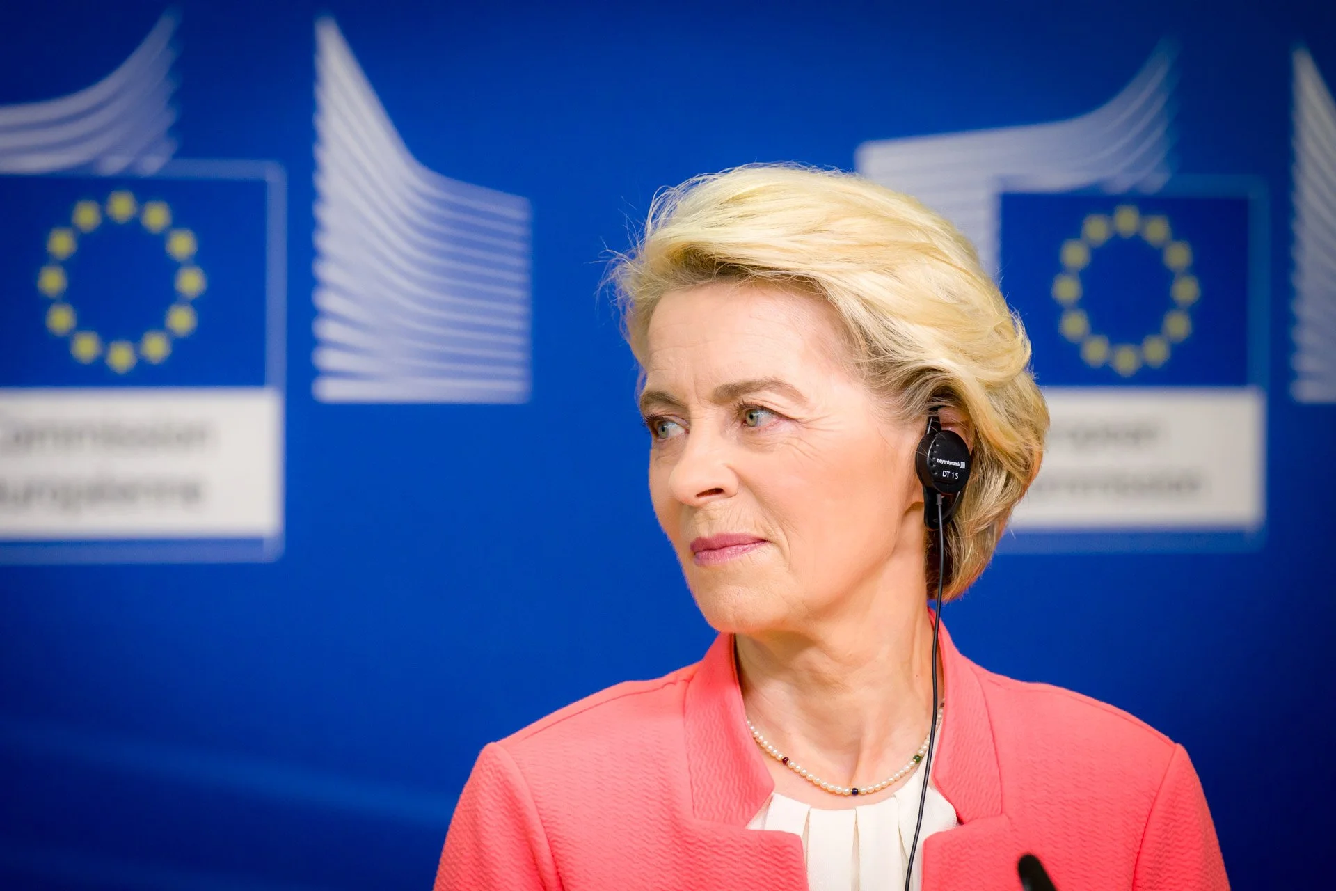 Une femme blonde avec un casque écouteur, vêtue d'une veste rose, devant un fond bleu avec le logo de l'Union Européenne et l'inscription "Commission Européenne".