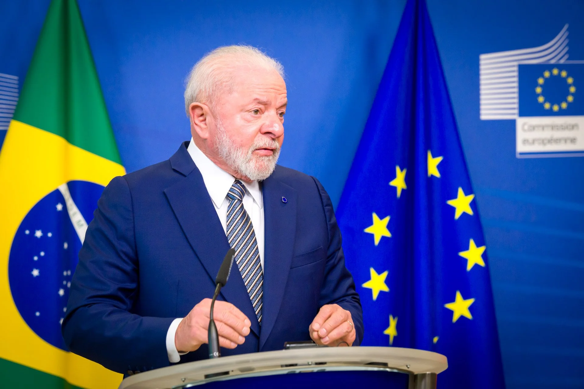 Un homme en costume parlant à un podium, avec les drapeaux du Brésil et de l'Union européenne en arrière-plan.