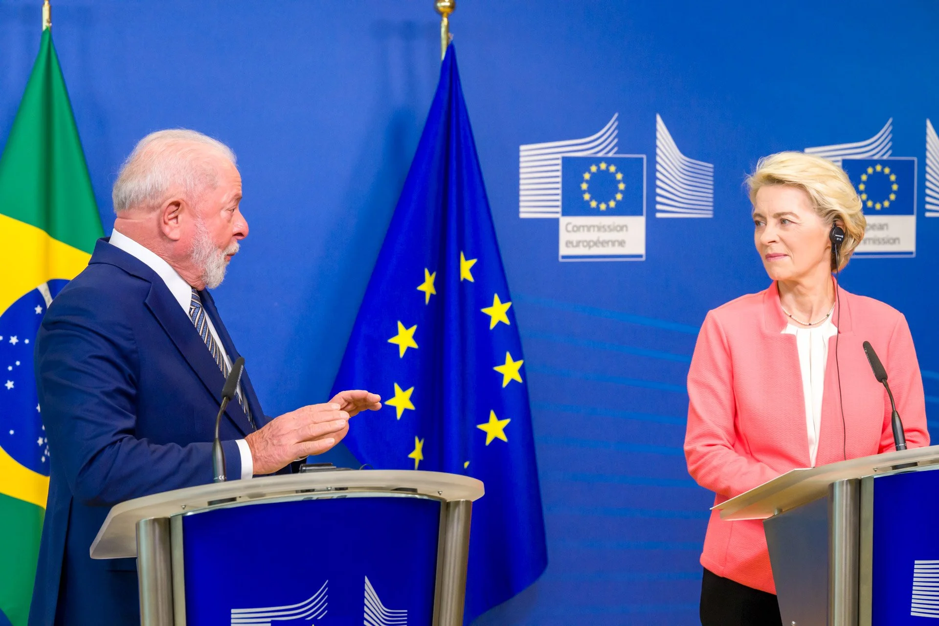 Deux personnes debout devant des pupitres avec les drapeaux du Brésil et de l'Union européenne en arrière-plan. Un homme à gauche parle gesticulant avec les mains, et une femme à droite écoute attentivement.