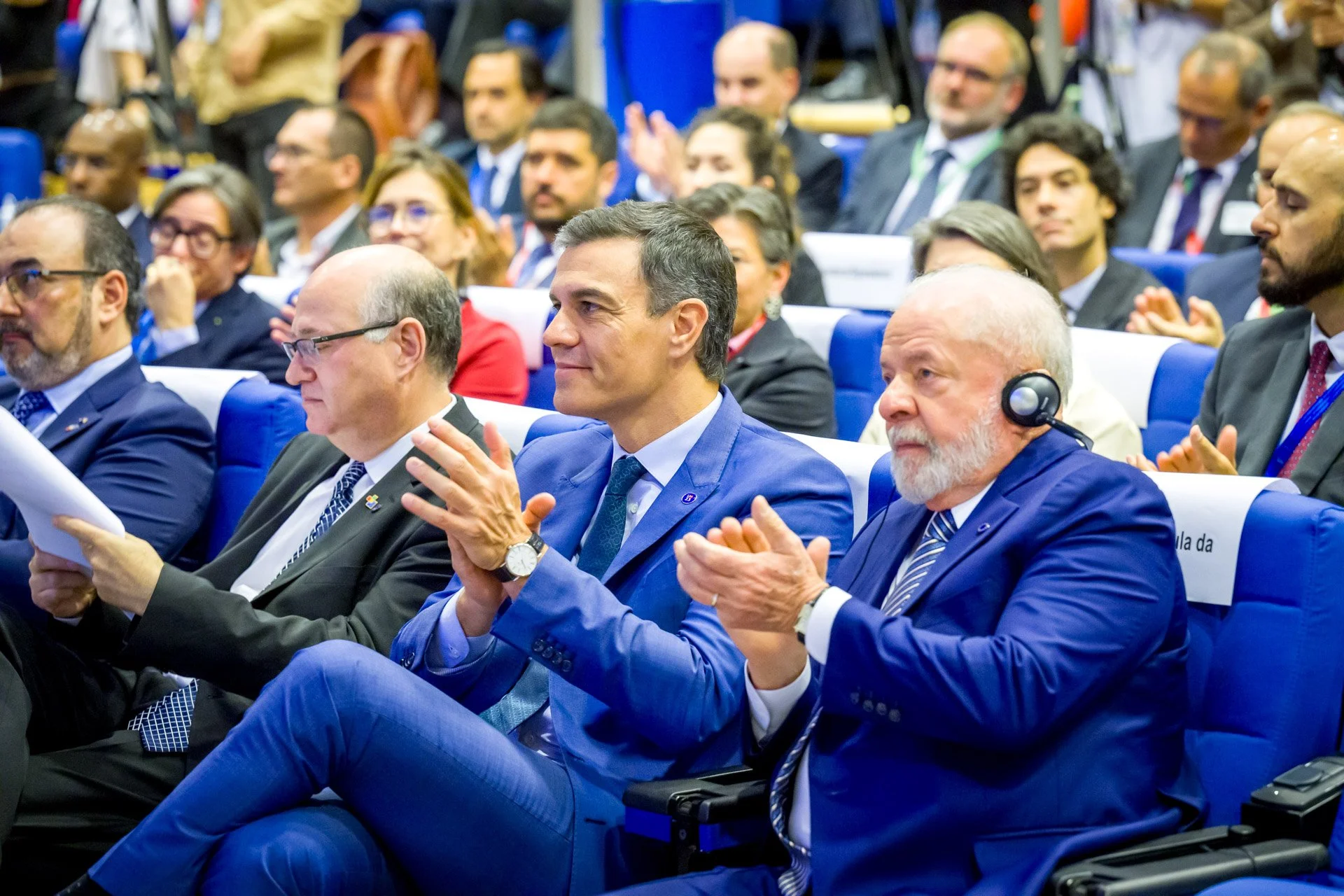 Une assemblée de personnes en costume assises, certaines applaudissant, dans ce qui semble être une conférence ou un événement officiel.