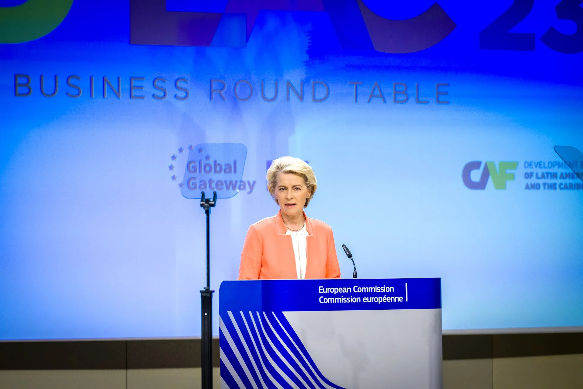 Femme en costume orange s'exprimant à un podium de la Commission européenne, avec les logos "Global Gateway" et "CAF" en arrière-plan.
