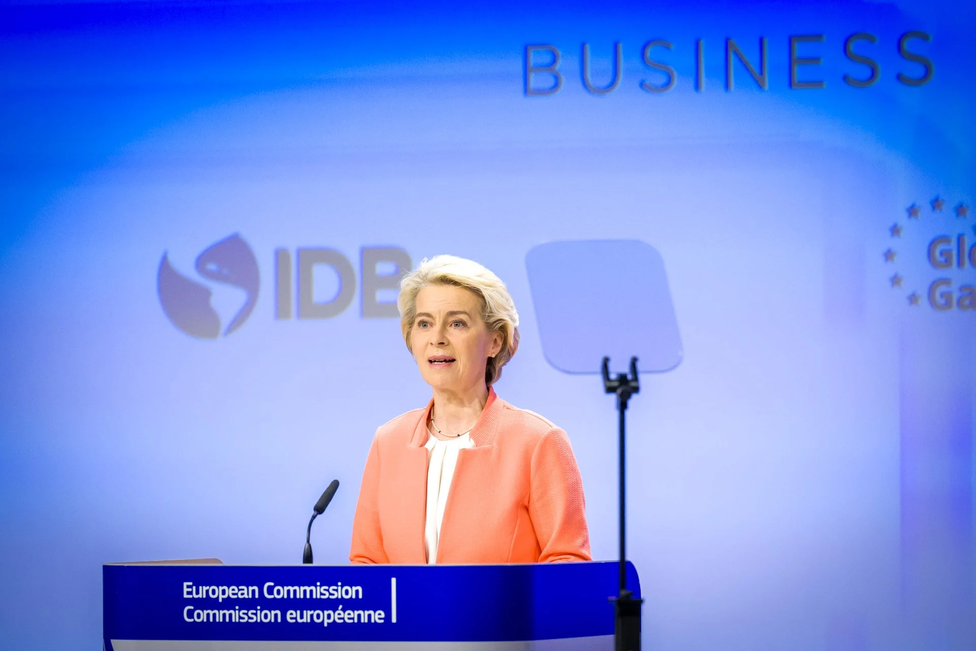 Femme en costume orange lors d'une conférence de presse de la Commission européenne, avec le logo de l'IDB en arrière-plan.