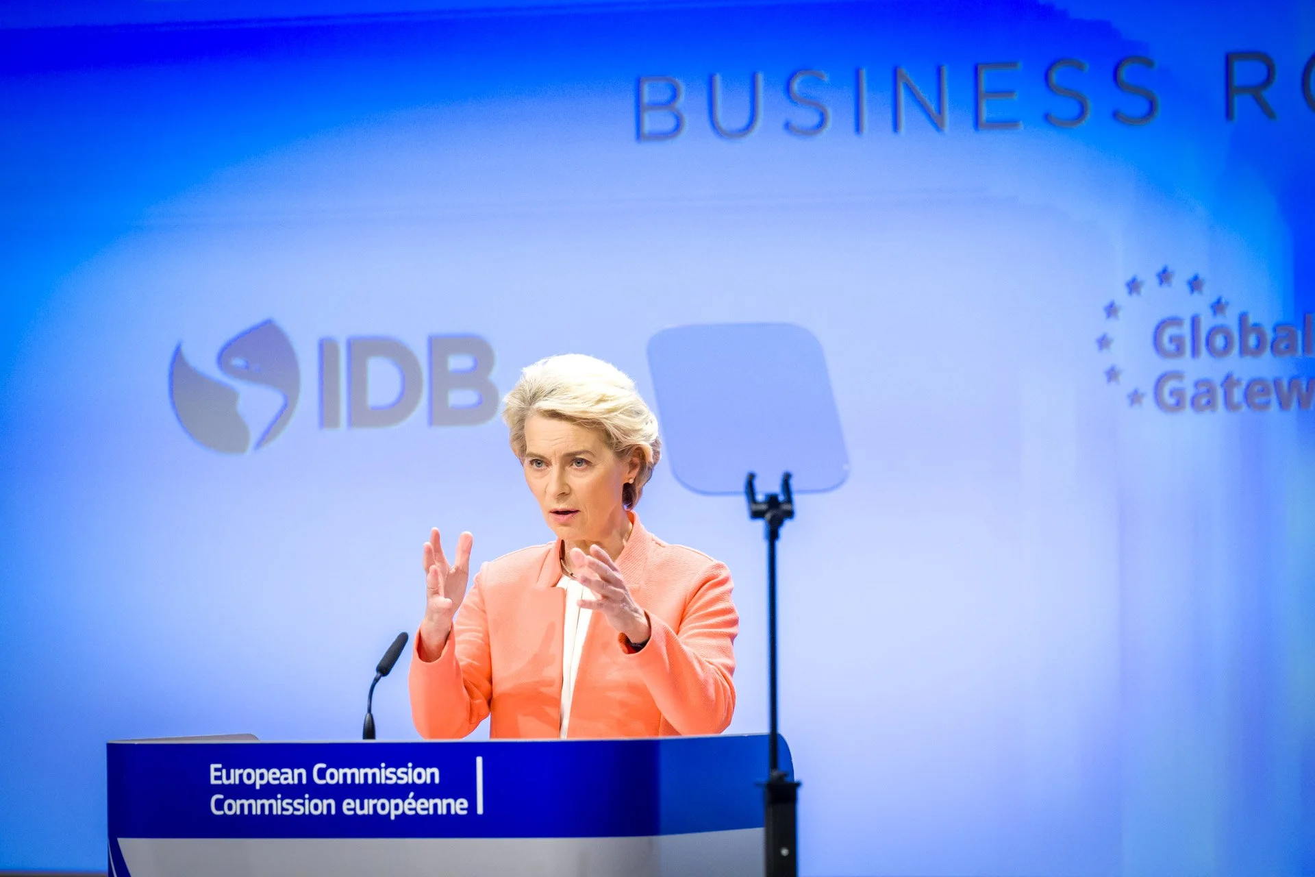 Une personne en costume rose parle à un podium avec le logo de la Commission européenne et IDB en arrière-plan.
