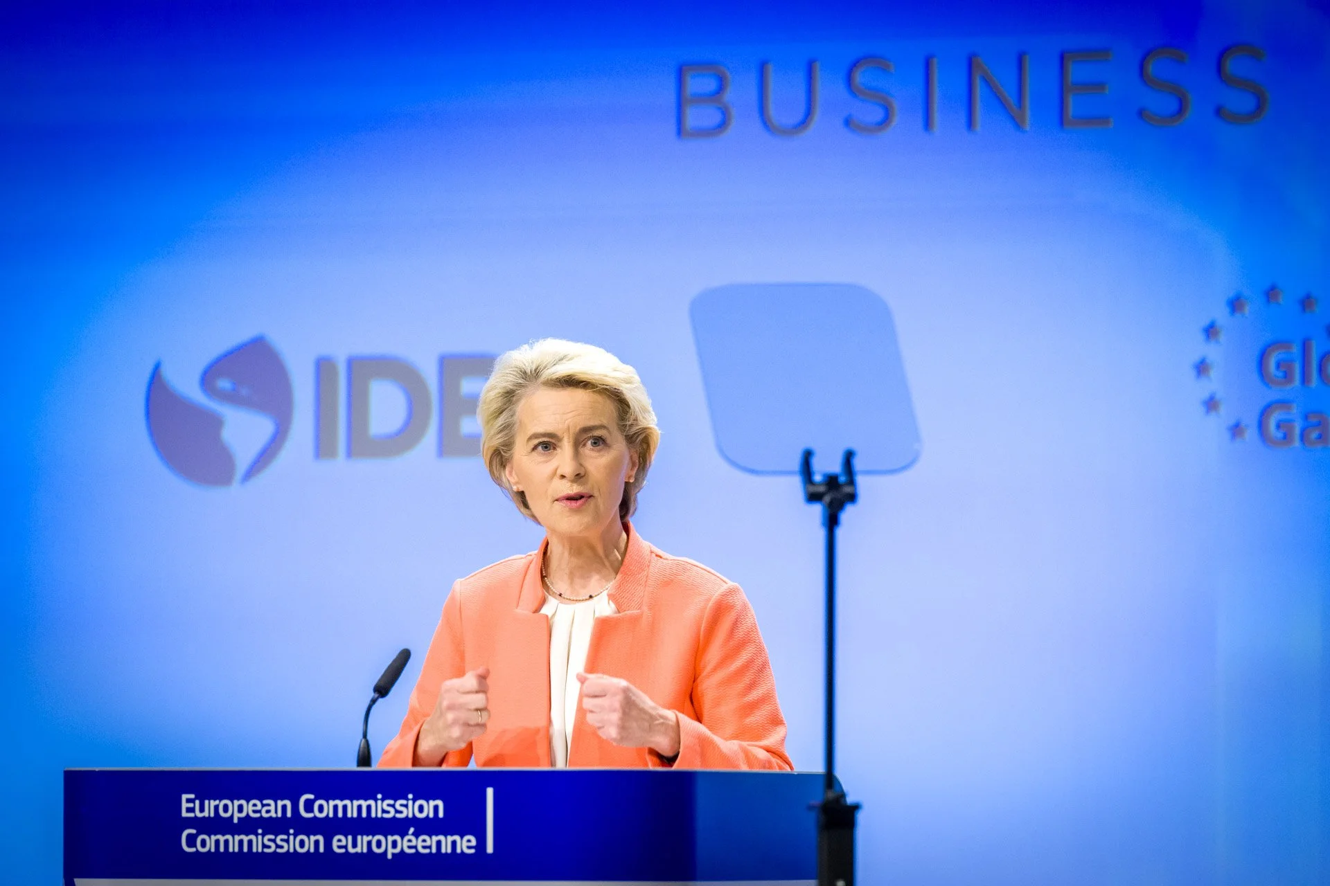 Une femme en veste orange parle à un pupitre marqué "Commission européenne". En arrière-plan, des logos et mots liés au business sont visibles.