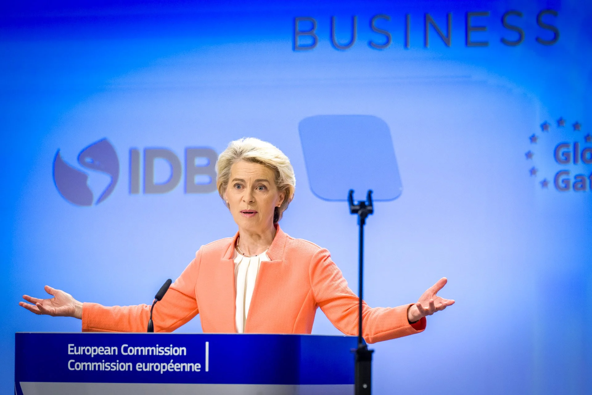 Personne en tailleur orange parlant lors d'une conférence avec un pupitre de la Commission européenne en arrière-plan.