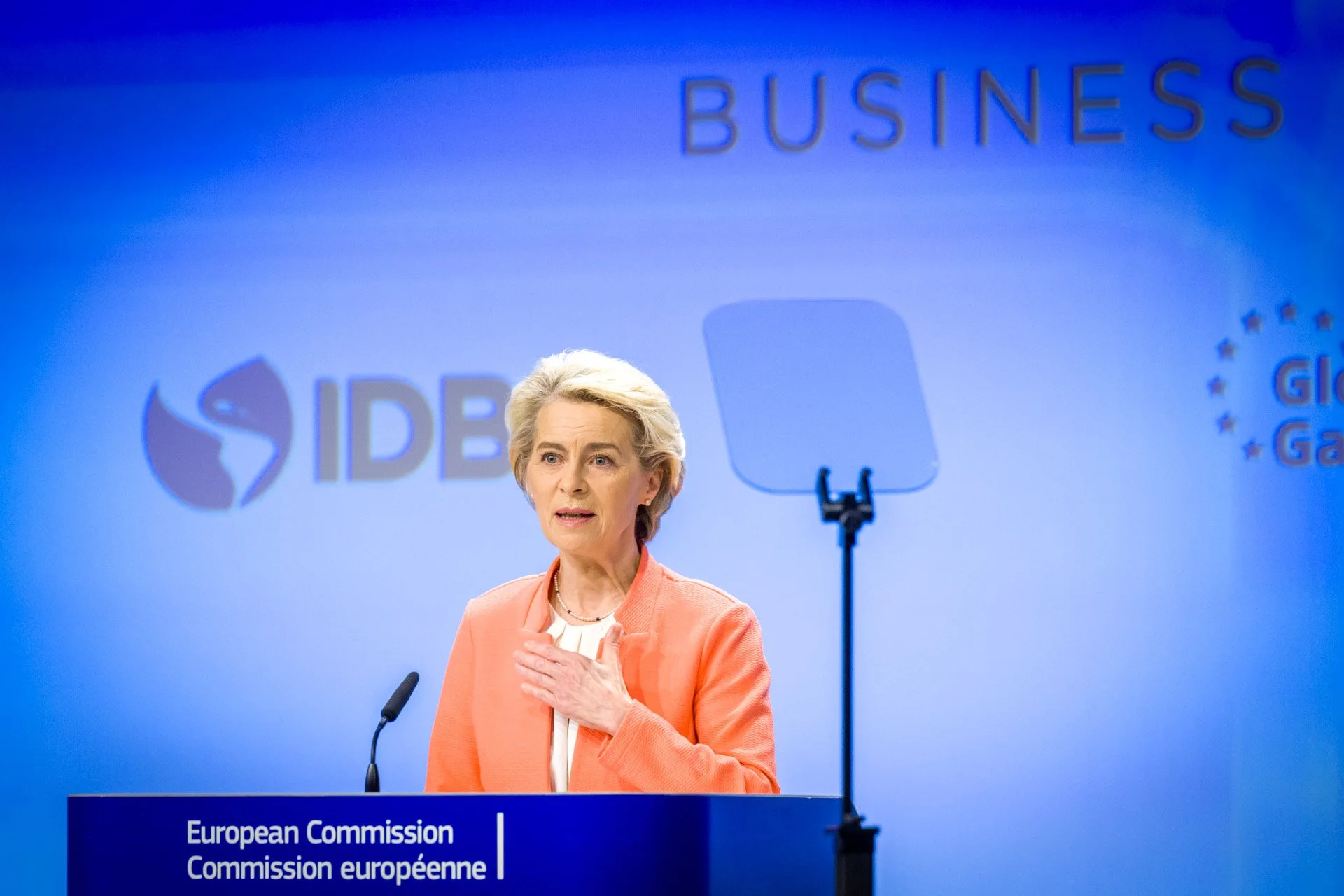 Une femme en discours devant un podium marqué 'Commission européenne' avec des logos à l'arrière-plan, sur un fond bleu.
