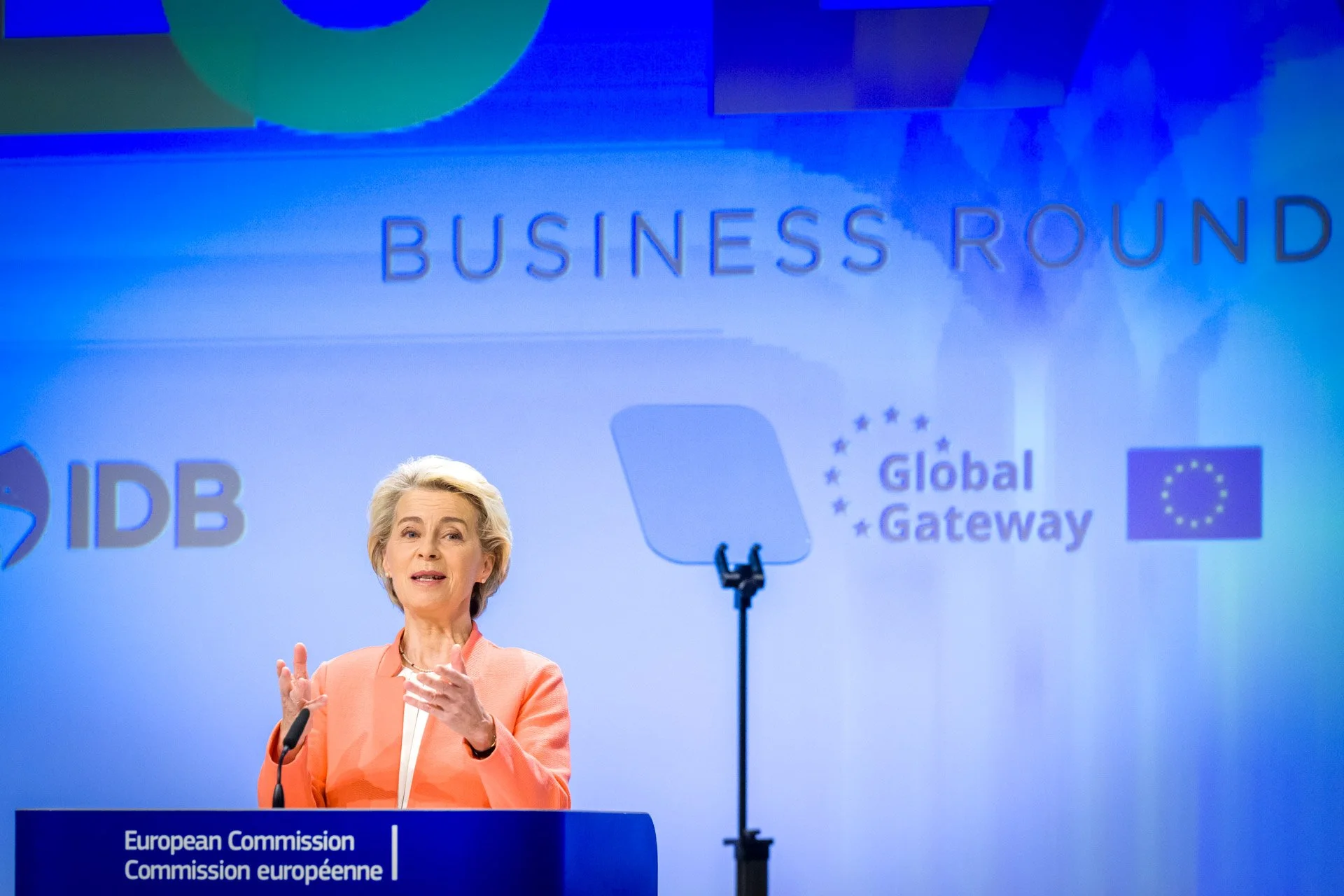 Femme en costume rose donnant un discours devant un podium de la Commission européenne, avec des logos IDB et Global Gateway en arrière-plan.