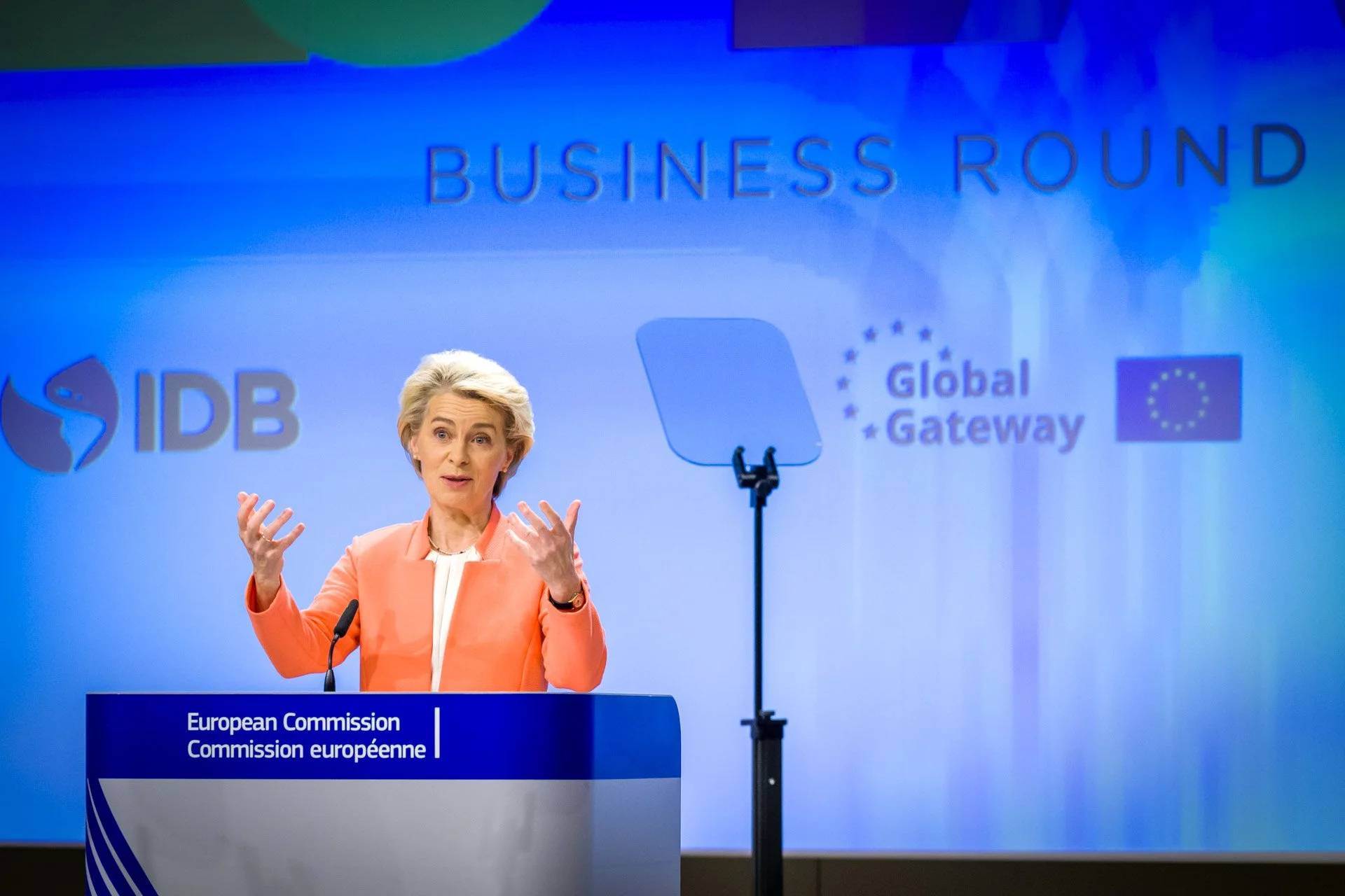 Une personne en veste orange parle devant un pupitre de la Commission européenne. Derrière, des logos "IDB" et "Global Gateway" sont visibles, avec une mention "BUSINESS ROUND" en haut.