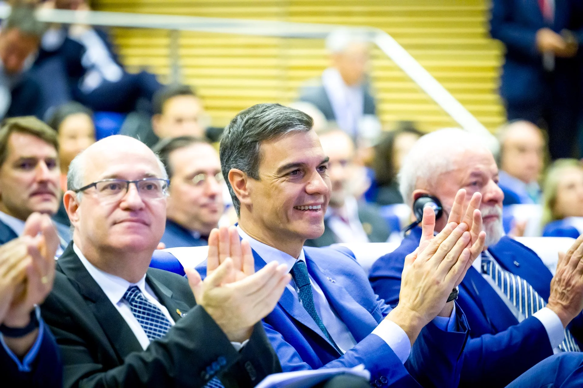 Homme en costume bleu applaudissant lors d'une conférence. Autres personnes autour, en arrière-plan flou.