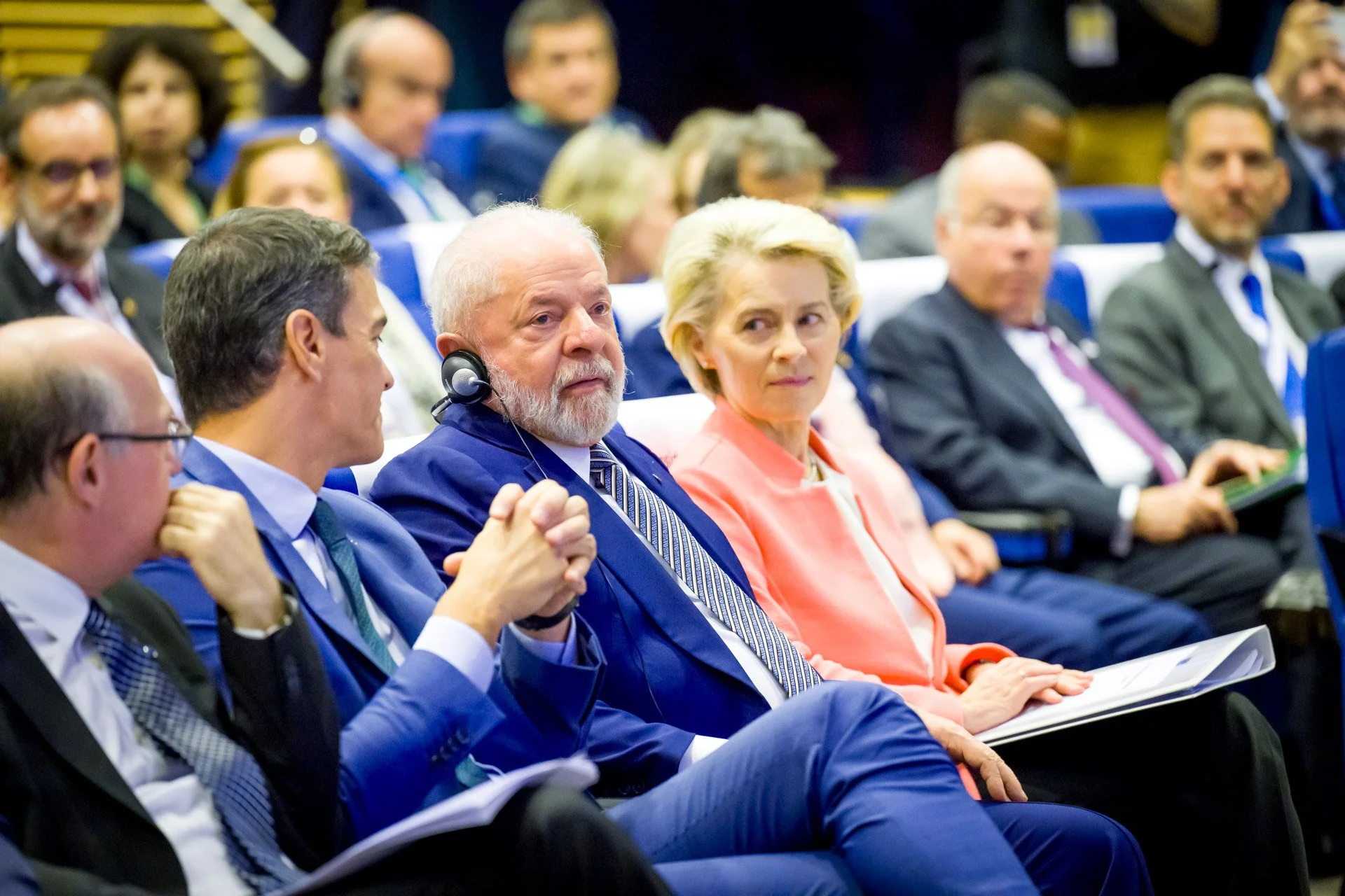 Des personnes en costume assises lors d'une conférence ou d'un événement politique, portant des écouteurs pour la traduction simultanée.