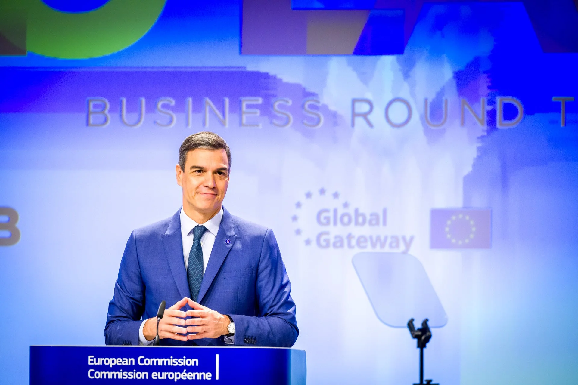 Un homme en costume bleu donne une présentation lors d'un événement avec un podium intitulé "European Commission" et le texte "Global Gateway" en arrière-plan.