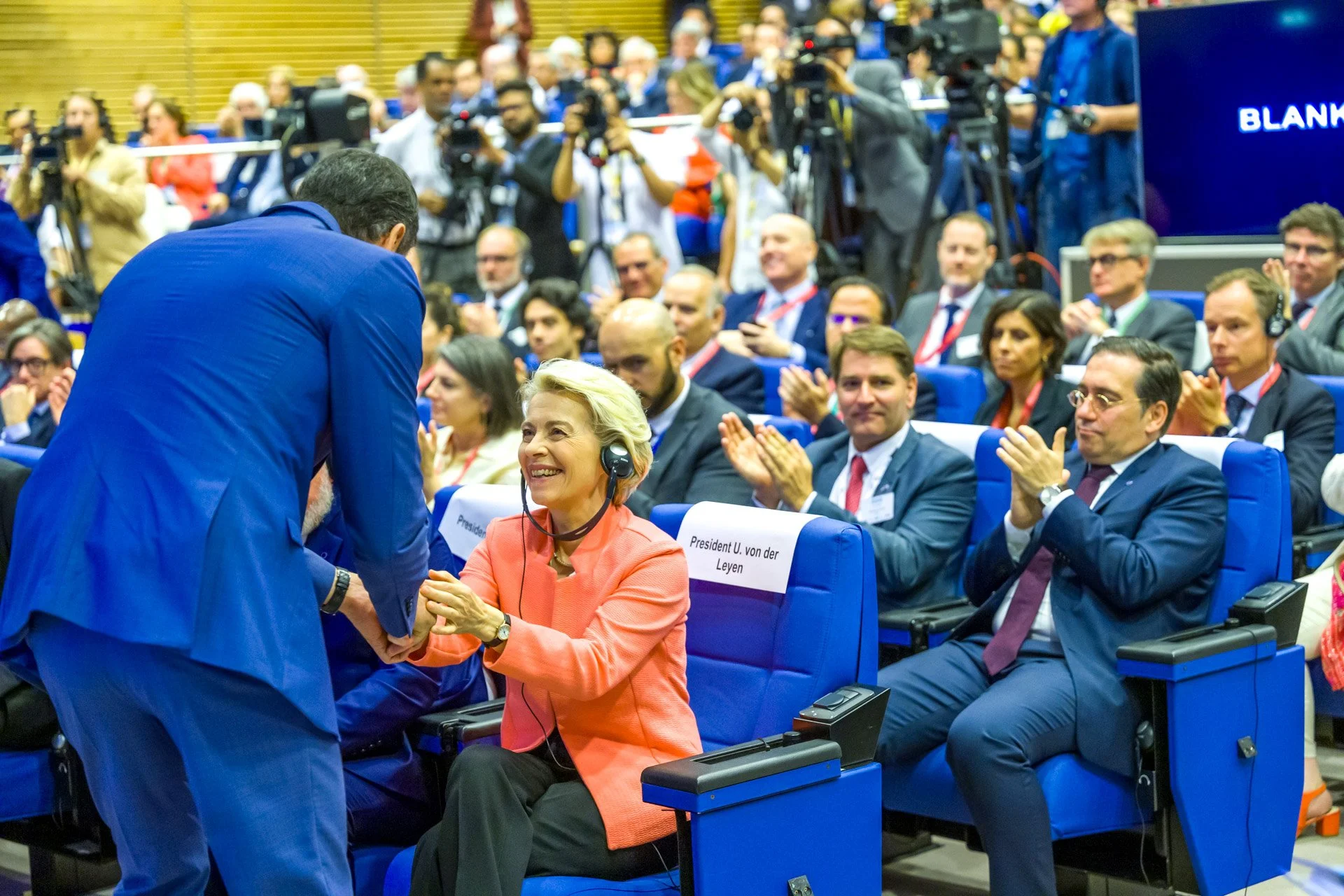 Personnes dans une conférence, une femme en rose serre la main d'un homme en costume bleu, entourées d'une audience avec des écouteurs, certaines applaudissent, et des caméras à l'arrière-plan.