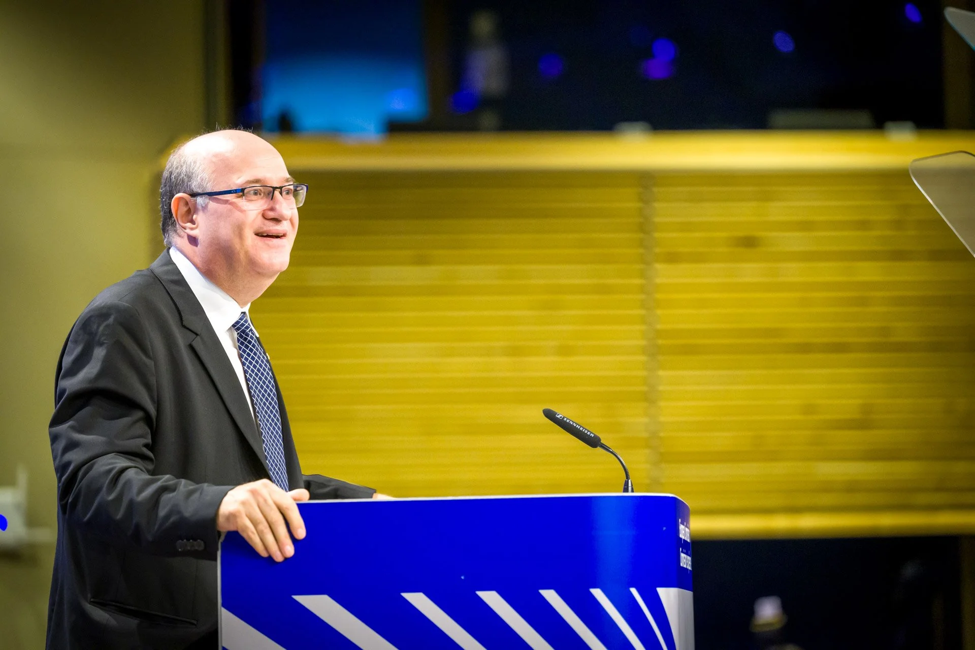 Un homme en costume souriant debout derrière un podium bleu avec un microphone.