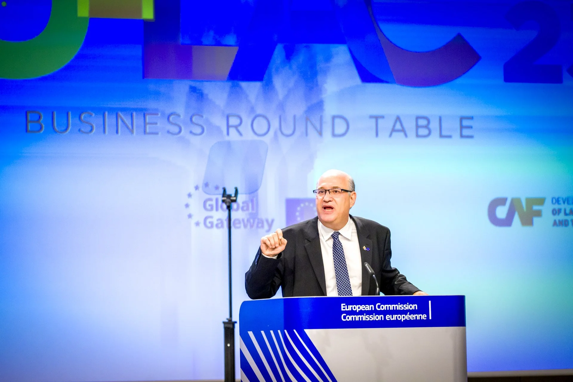 Un homme en costume parle sur un podium lors d'une conférence autour d'une table ronde sur les affaires, organisé par la Commission européenne. En arrière-plan, on voit les mots "Global Gateway" et des logos institutionnels.