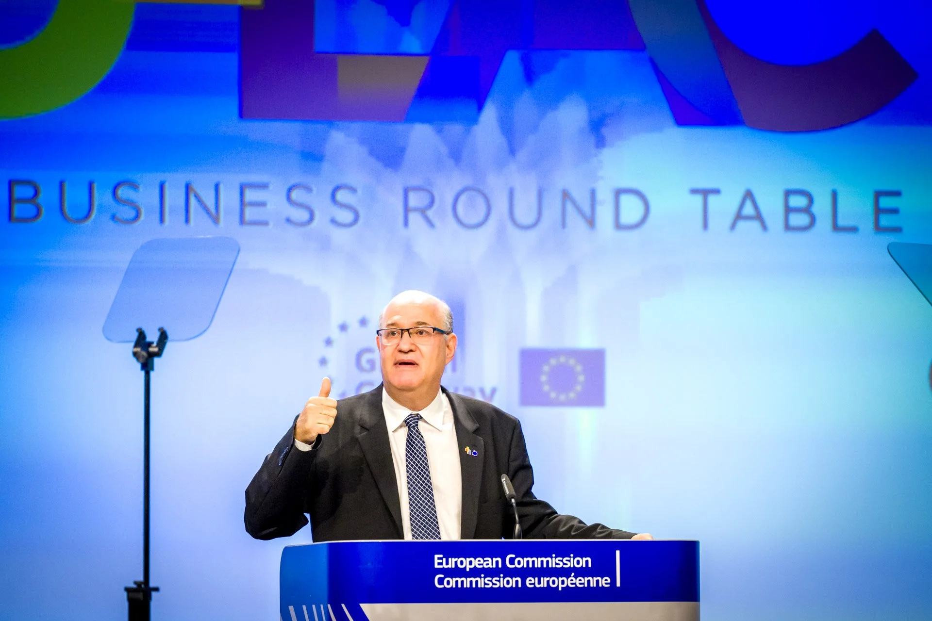Un homme en costume parle à un podium de la Commission européenne lors d'une table ronde sur les affaires. Un écran avec le texte 'Business Round Table' est visible en arrière-plan.