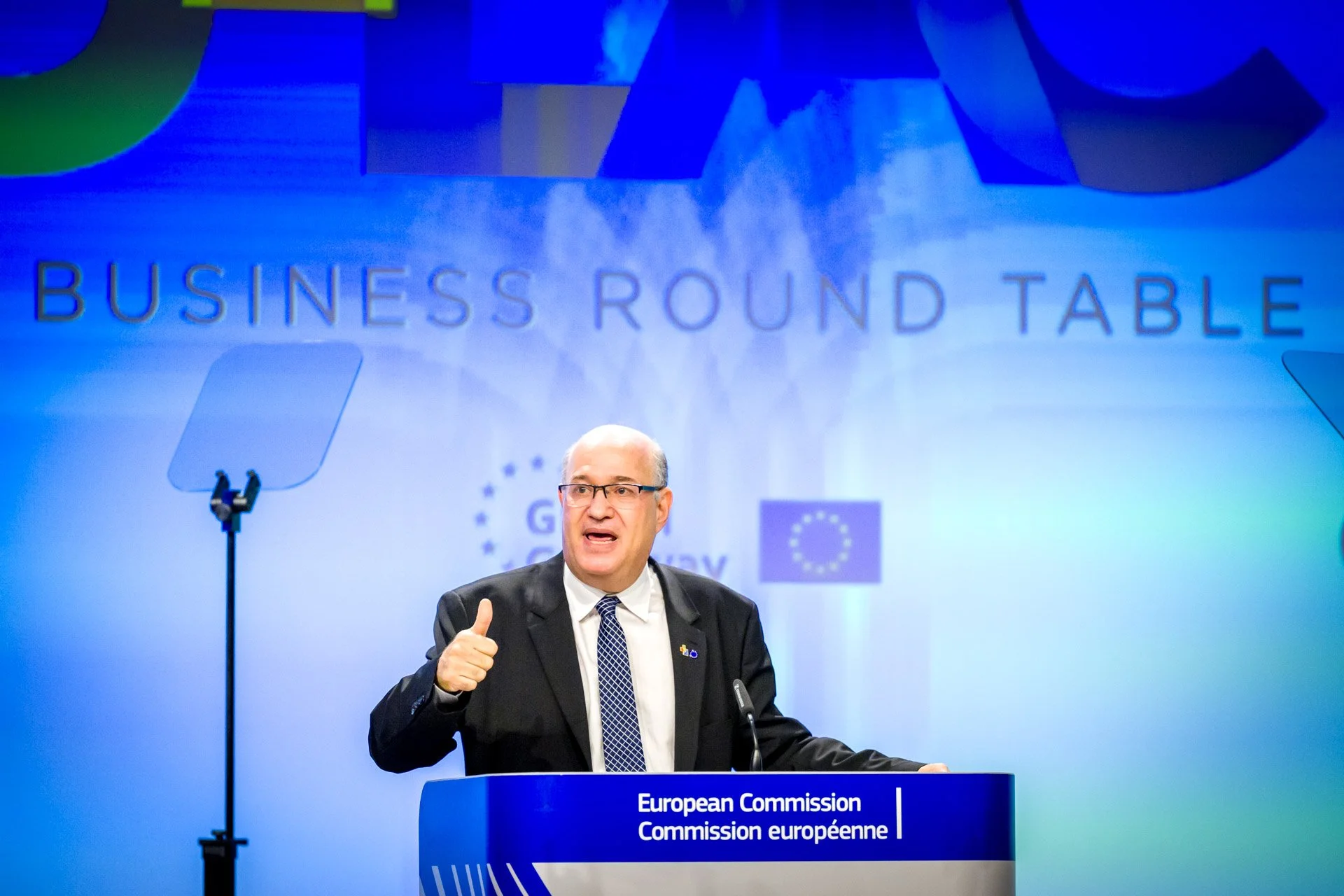 Un homme en costume donne un discours à une tribune avec l'inscription "Commission européenne". Le fond est bleu avec le texte "BUSINESS ROUND TABLE". Un écran de prompteur est visible.