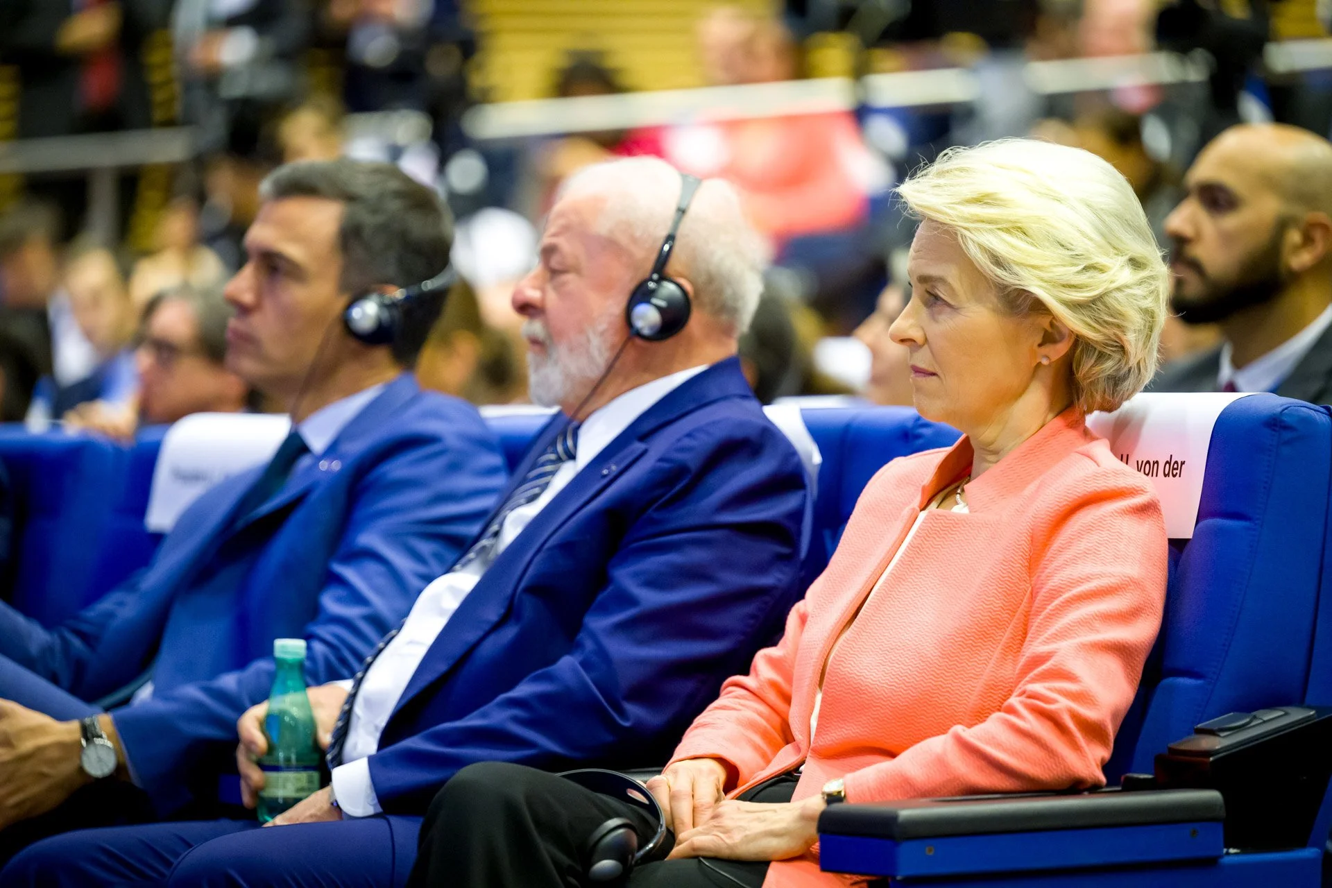 Des personnes assises à une conférence, portant des écouteurs avec des micros. Au premier plan, une femme portant un blazer orange est concentrée. Les sièges sont bleus et l'environnement semble formel.