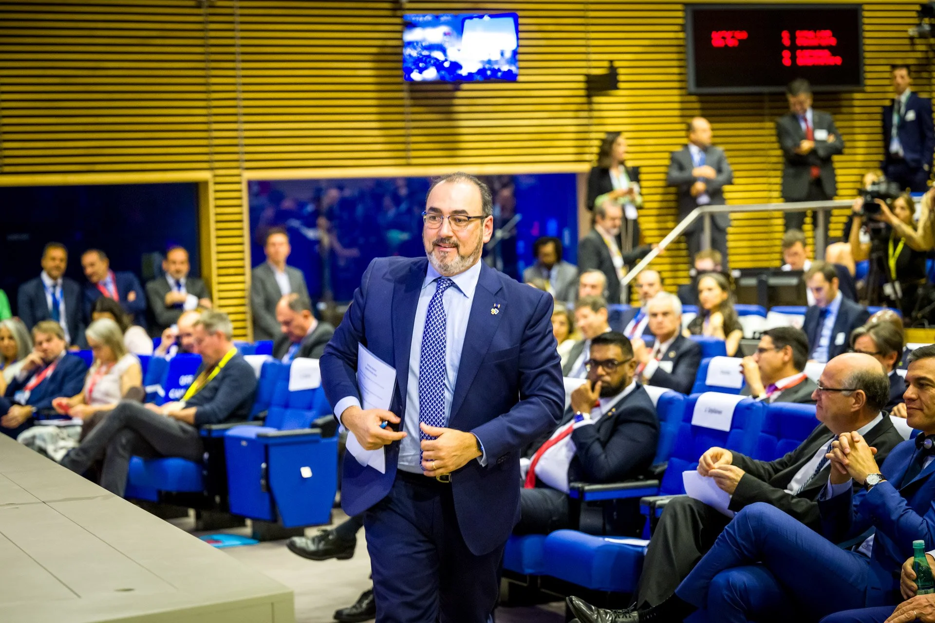 Un homme en costume marche dans une salle de conférence remplie de personnes assises sur des sièges bleus. Il tient des documents dans les mains et semble confiant. D'autres participants, également en tenue formelle, sont attentifs et assis autour de