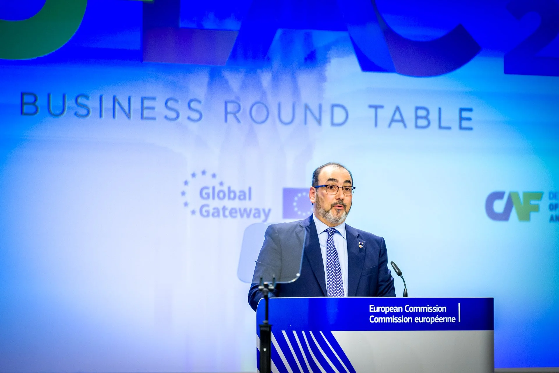 Homme en costume parlant derrière un podium lors d'une conférence de la Commission européenne, avec le logo "Global Gateway" en arrière-plan.