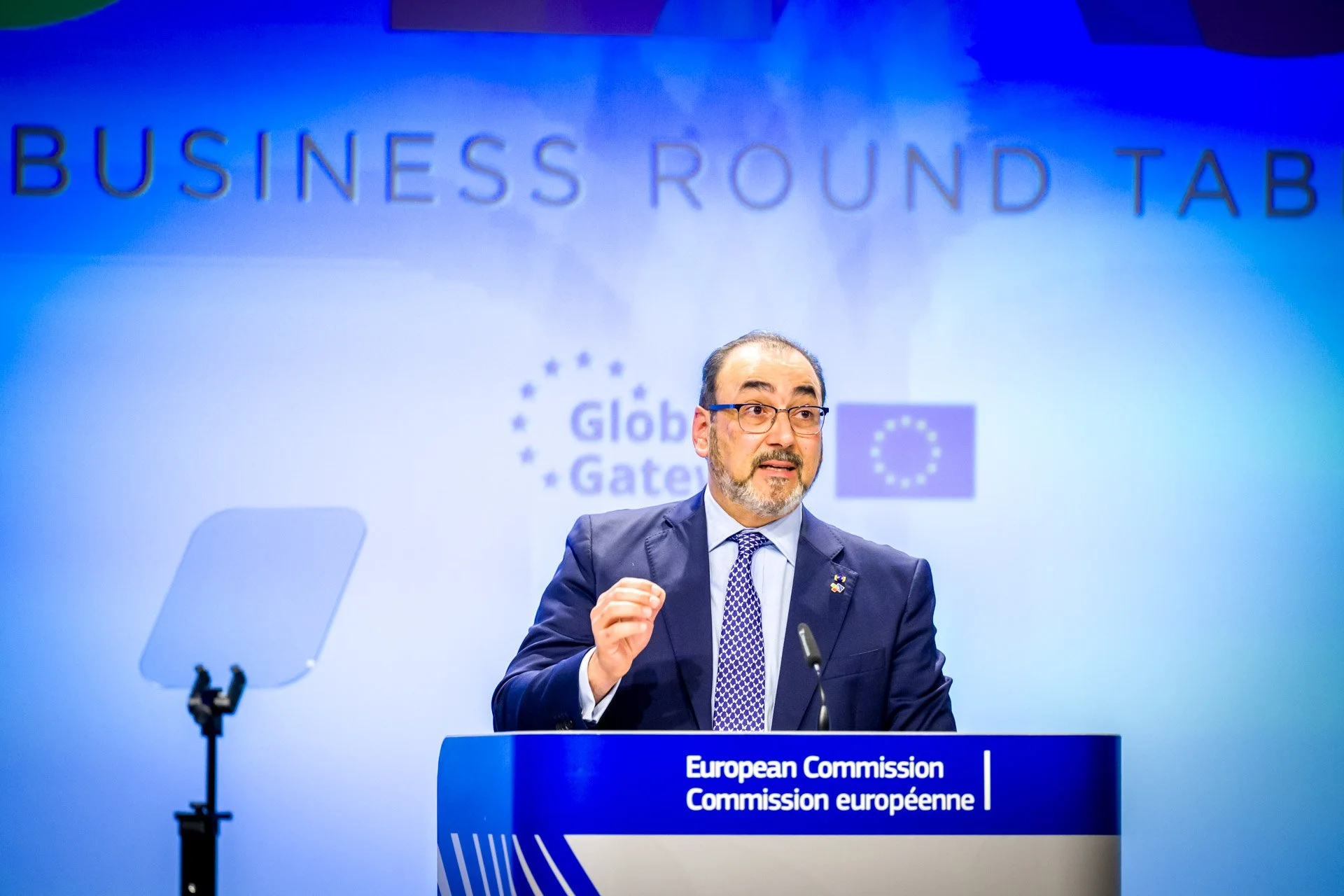 Un homme en costume parle lors d'une conférence de la Commission européenne. Il est debout devant un pupitre avec les logos de l'Union européenne en arrière-plan.