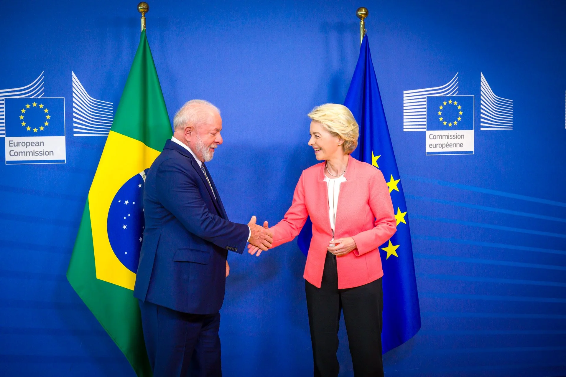 Deux personnes se serrent la main devant les drapeaux du Brésil et de l'Union européenne, avec le logo de la Commission européenne en arrière-plan.