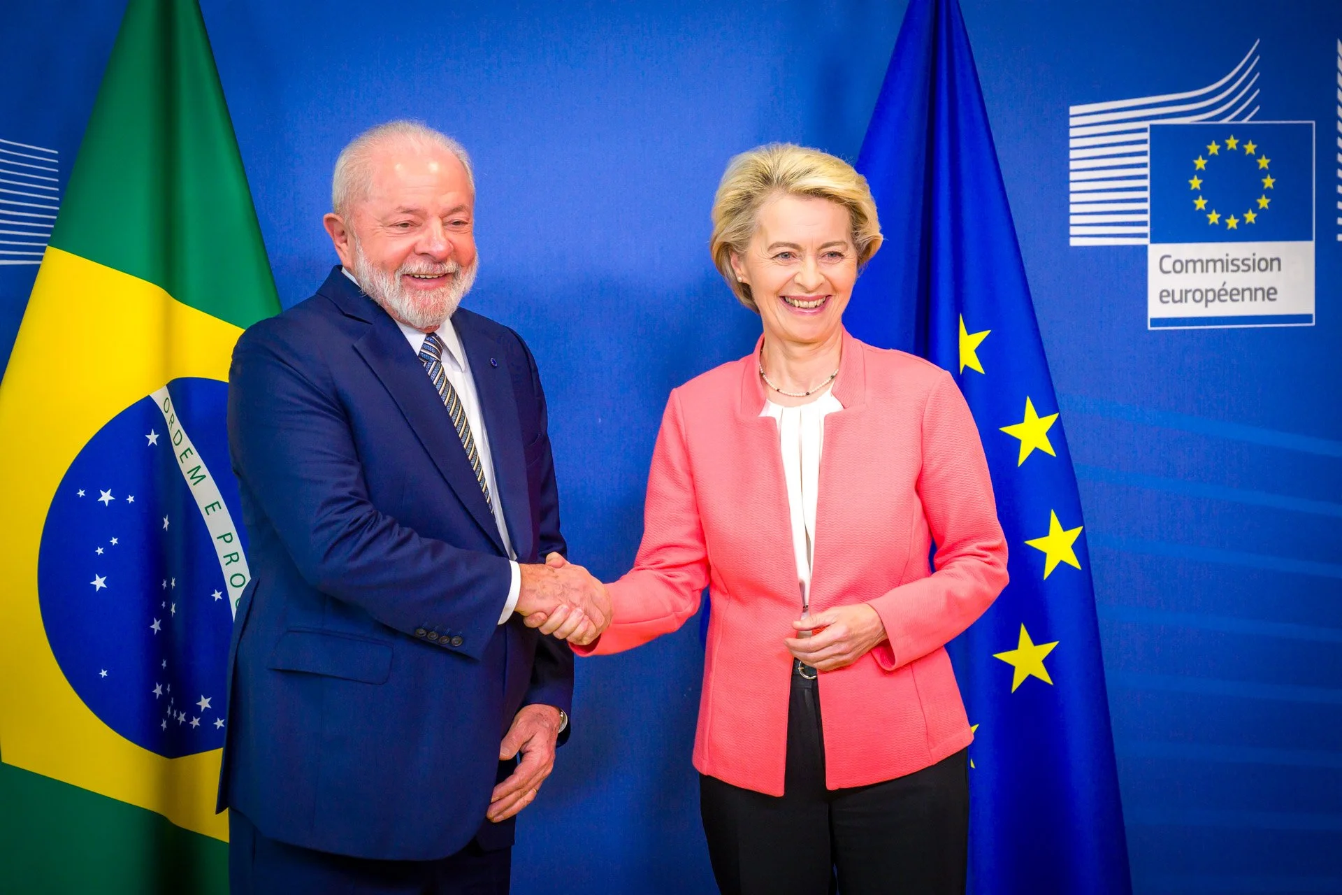 Deux personnes se serrent la main devant les drapeaux du Brésil et de l'Union européenne, avec un panneau de la Commission européenne en arrière-plan.
