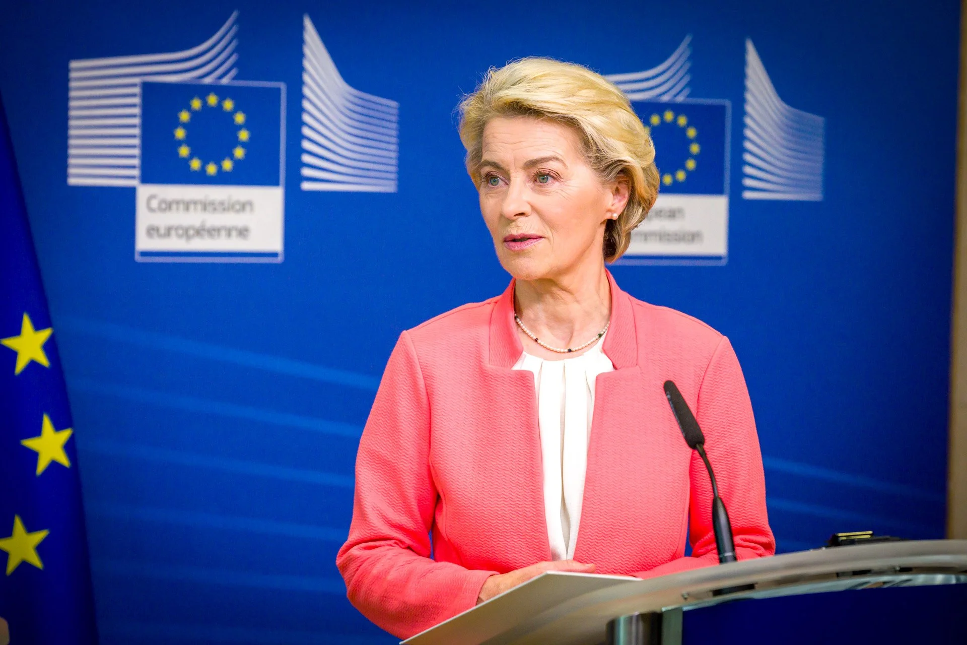Femme en veste rose, debout devant un podium, avec le drapeau de l'Union européenne en arrière-plan.