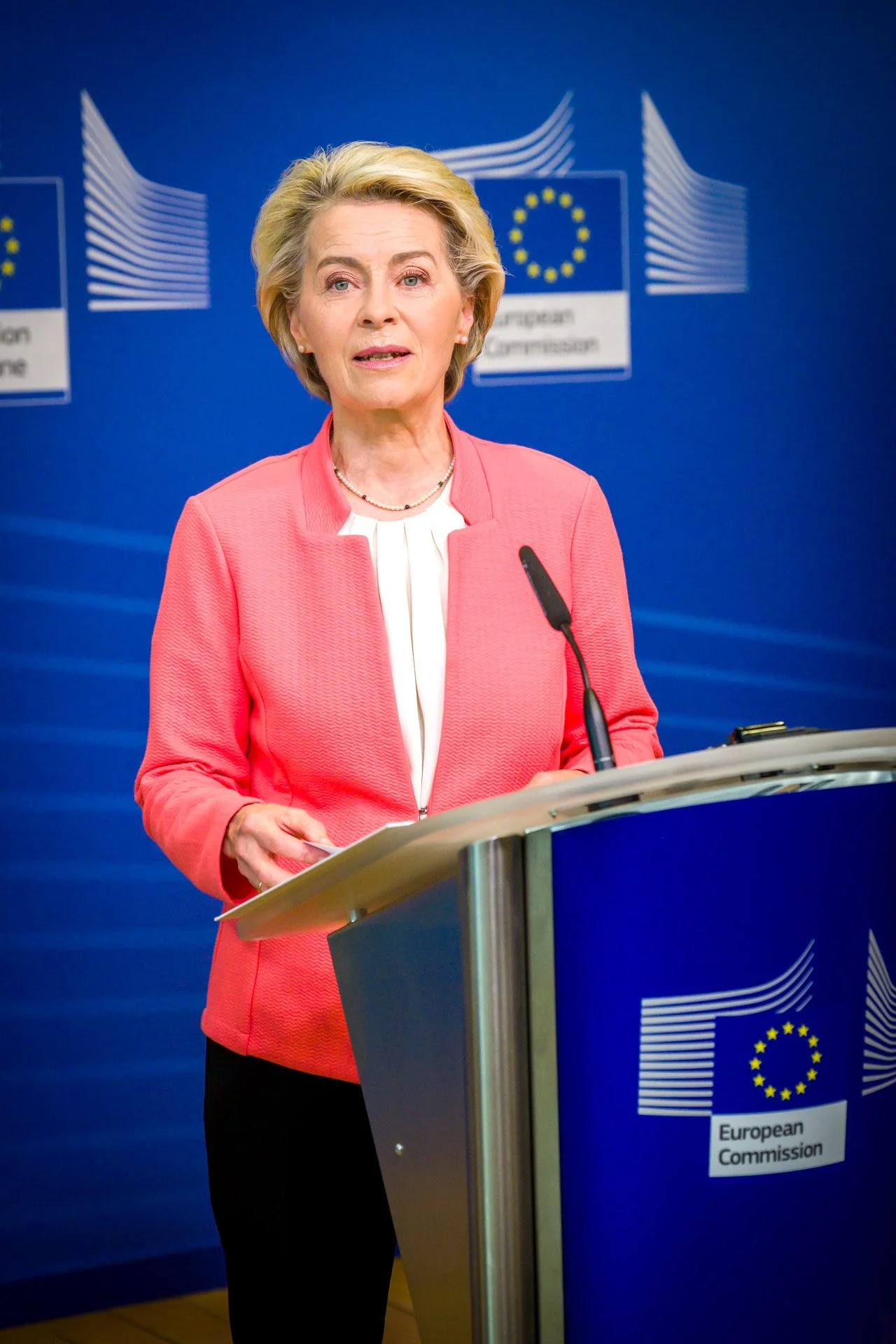 Femme en costume rose parlant à un podium avec emblèmes de la Commission européenne en arrière-plan.