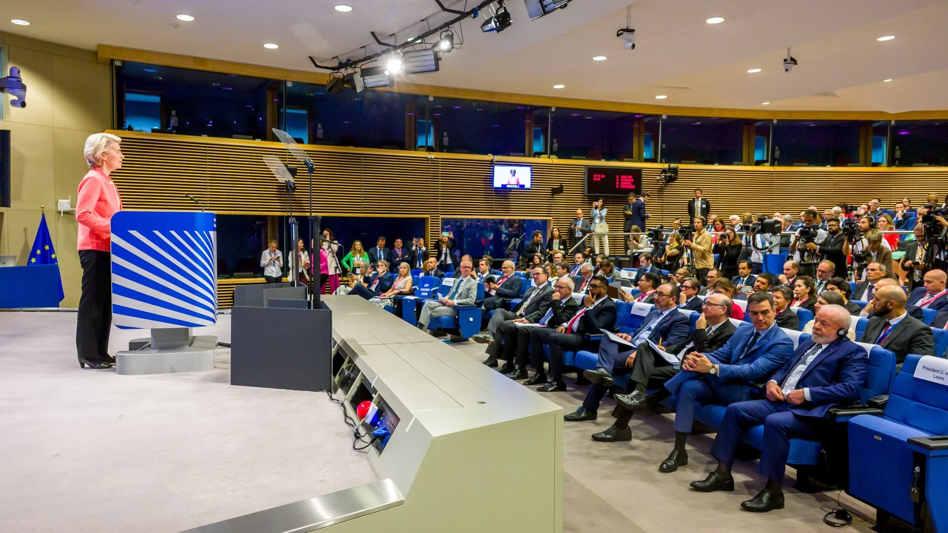 Conférence de presse avec une femme à un podium devant un public assis dans une salle moderne, drapeau de l'Union européenne en arrière-plan.