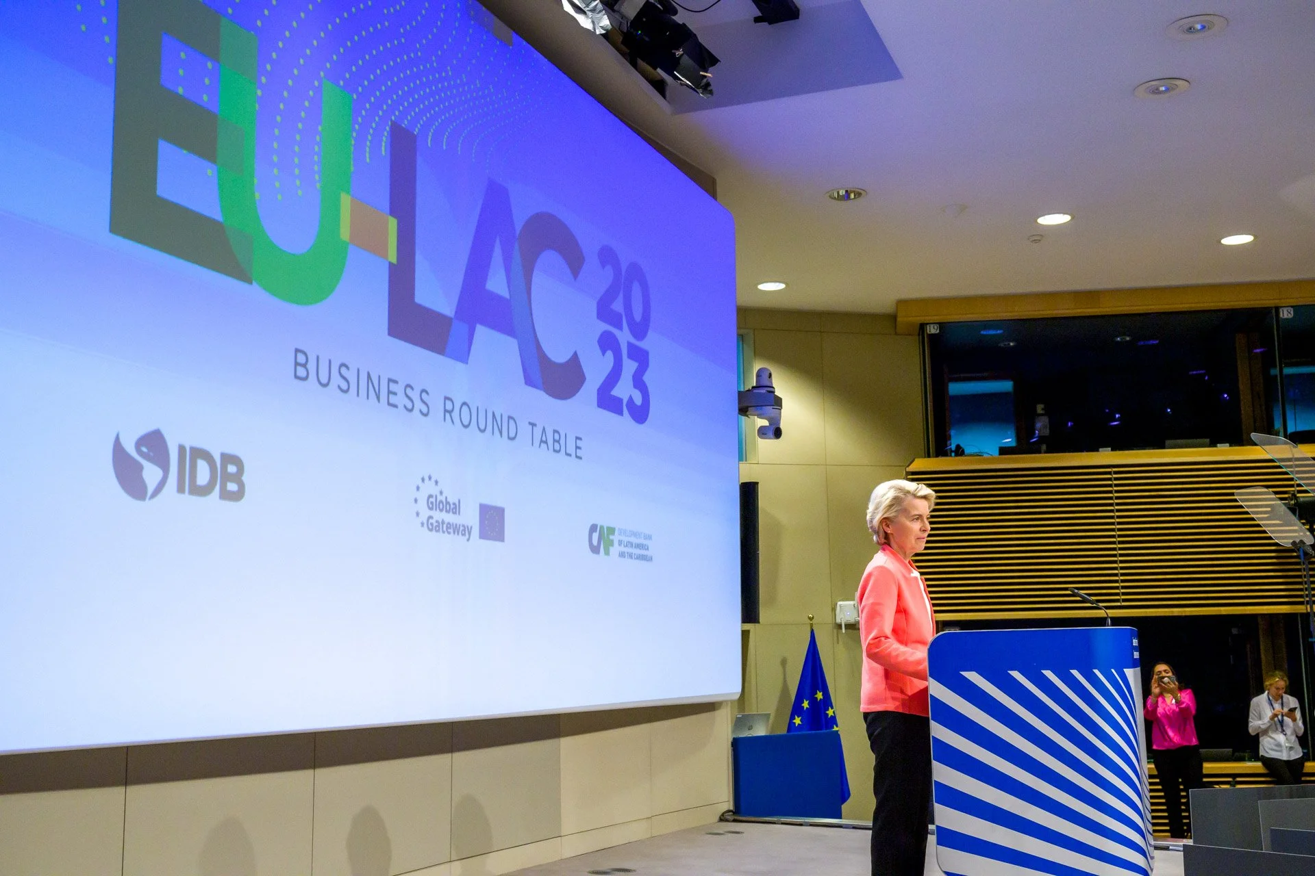 Une conférence avec le logo « EU-LAC 2023 Business Round Table » sur un écran, une personne en veste rose à un podium, et un drapeau de l'Union Européenne en arrière-plan.