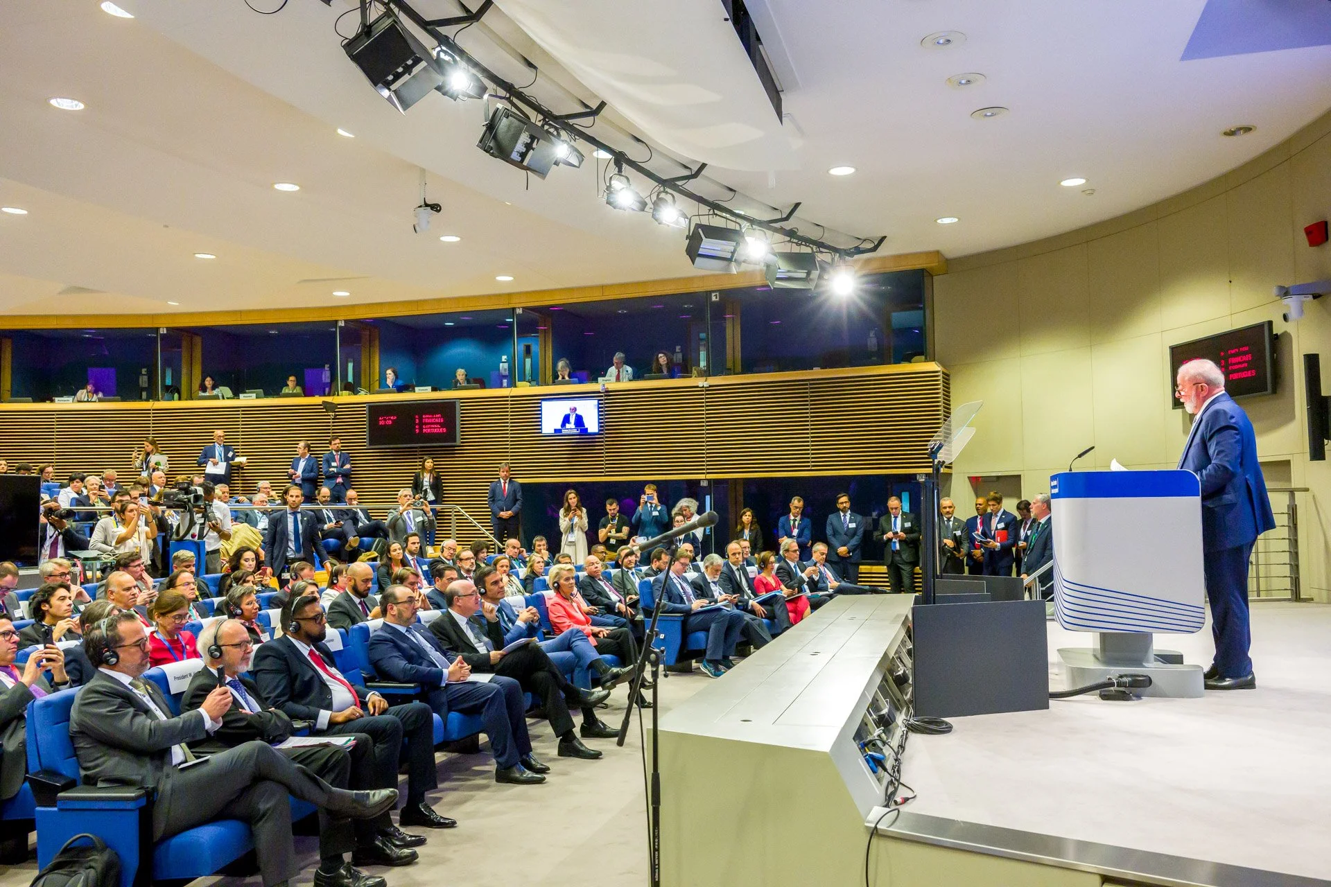 Un homme en costume fait un discours devant un public assis dans une salle de conférence moderne, avec des caméras et des personnes debout en arrière-plan.