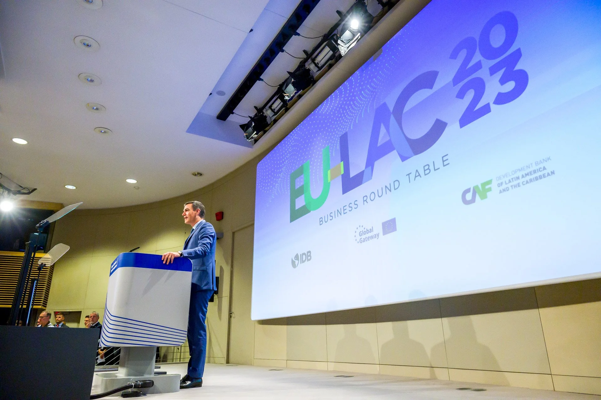 Conférence avec un orateur en costume bleu au pupitre devant un écran affichant "EU-LAC 2023 Business Round Table". Logos visibles incluent IDB, Global Gateway, et CAF. Salle de conférence avec public."