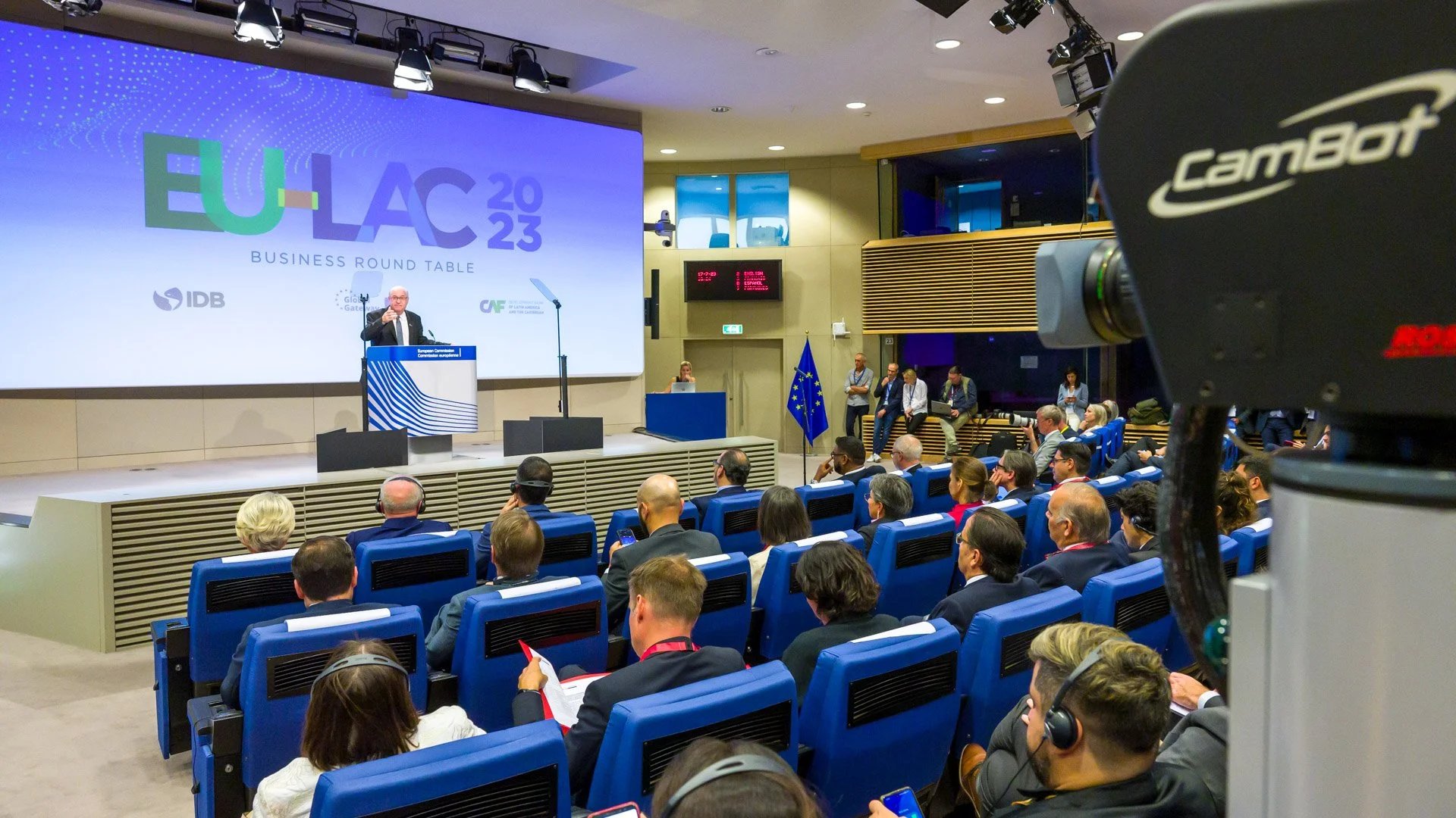 Conférence "EU-LAC 2023 Business Round Table" avec de nombreuses personnes assises et un orateur sur scène. Un écran affiche le logo de l'événement et une caméra est visible à l'avant-plan.