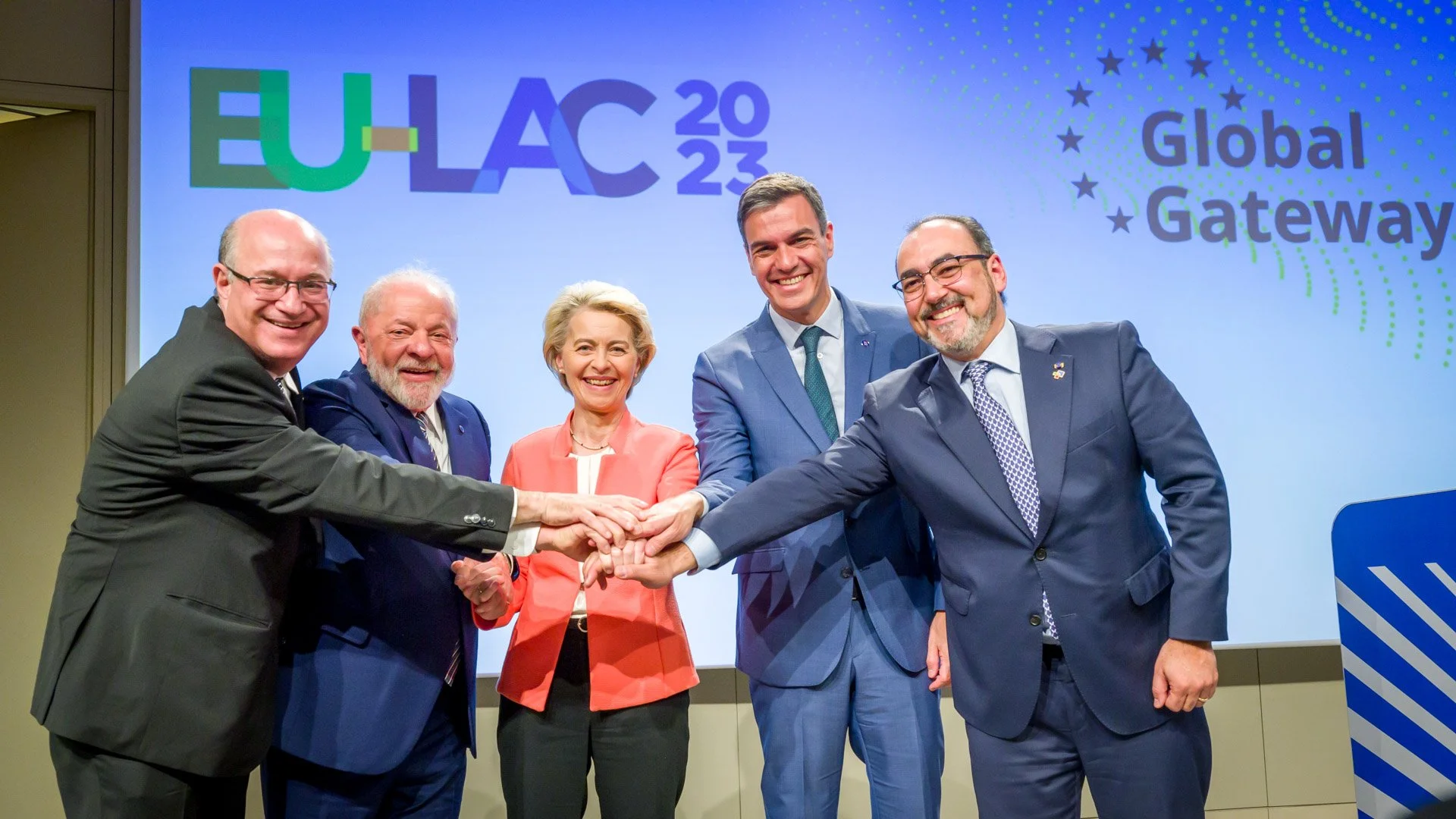 Cinq personnes en costume se tenant par la main devant un panneau "EU-LAC 2023" et "Global Gateway".