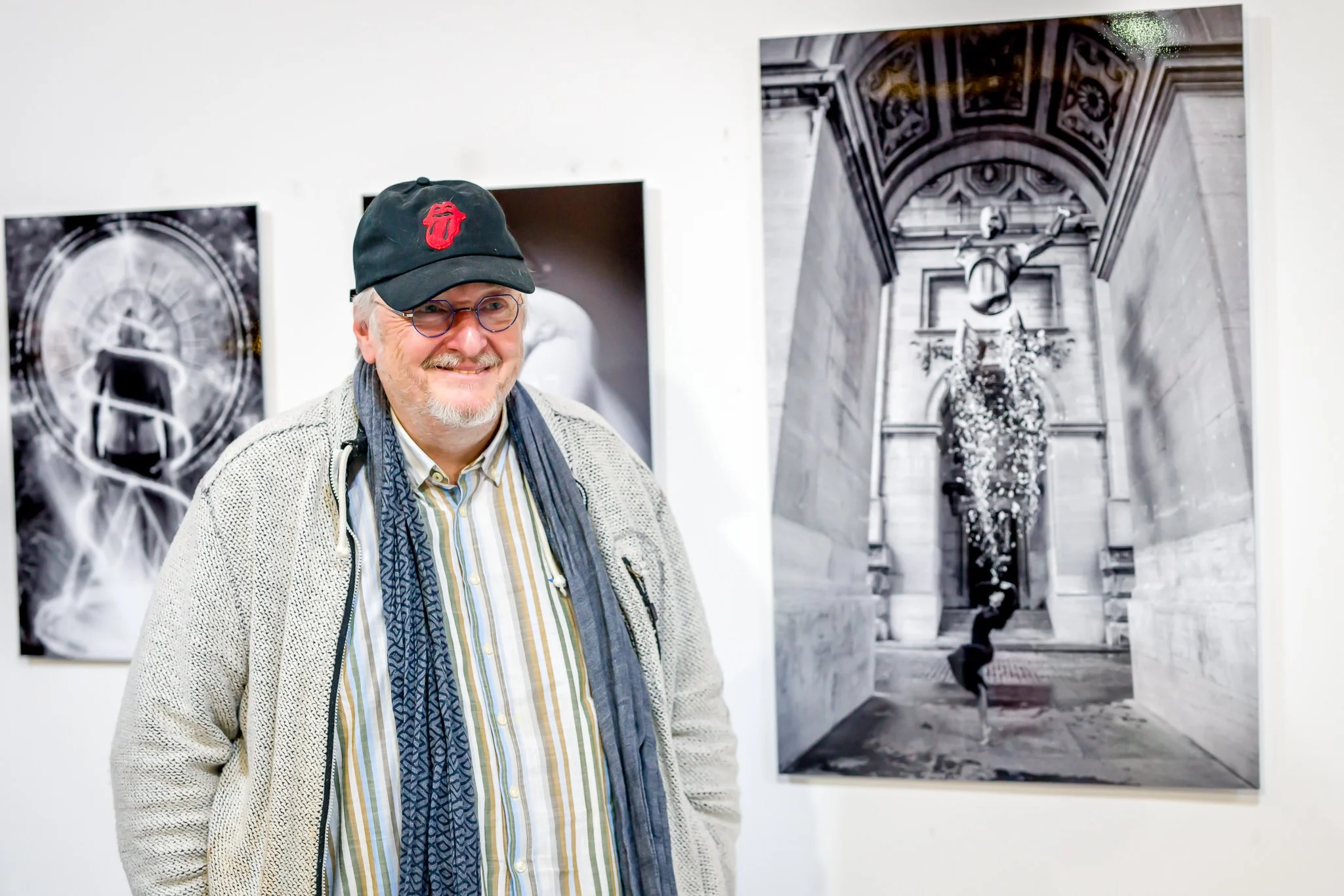 Un homme souriant portant une casquette et des lunettes se tient devant des photographies en noir et blanc exposées sur un mur.