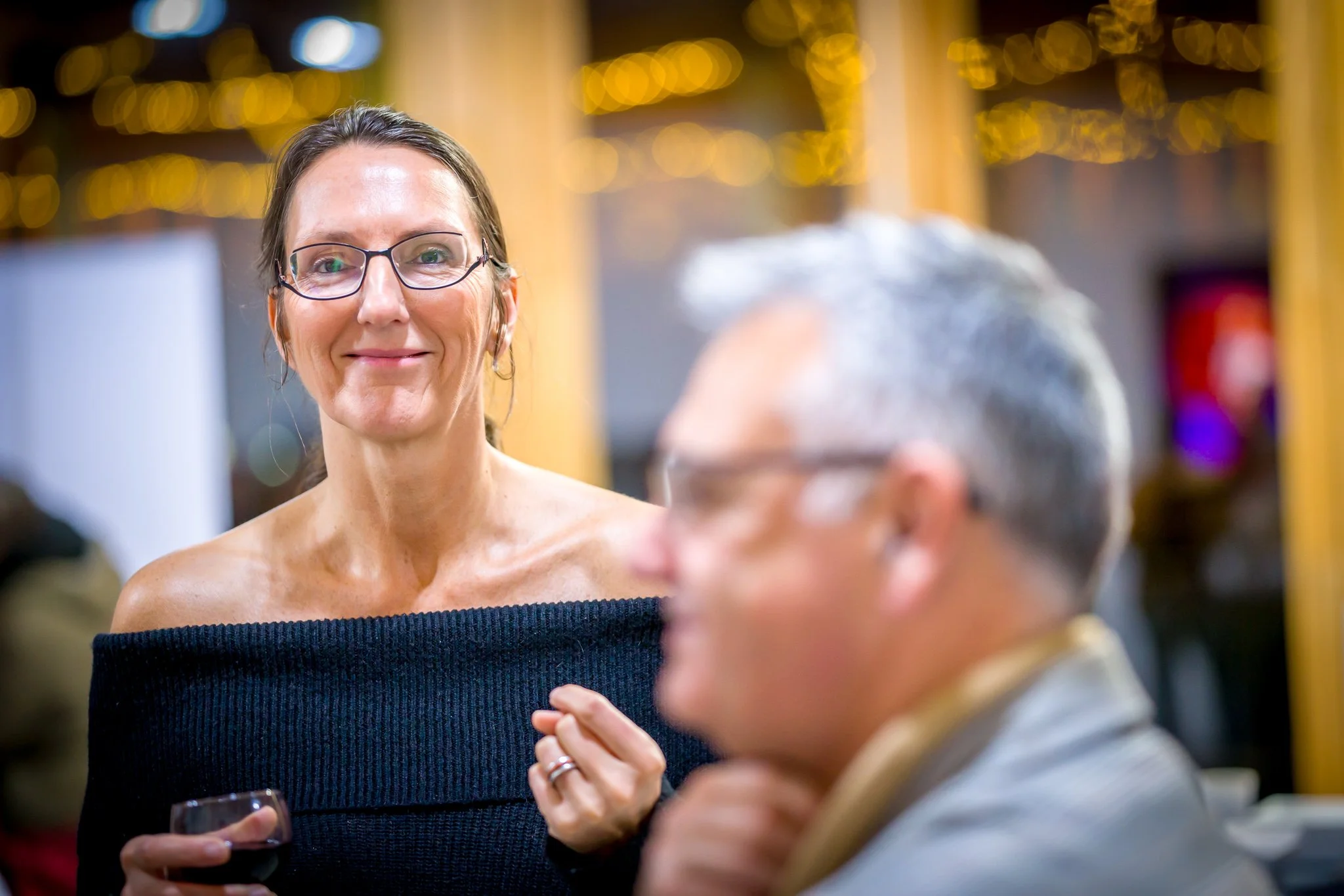 Une femme souriante avec des lunettes, tenant un verre, à côté d'un homme flou en arrière-plan.