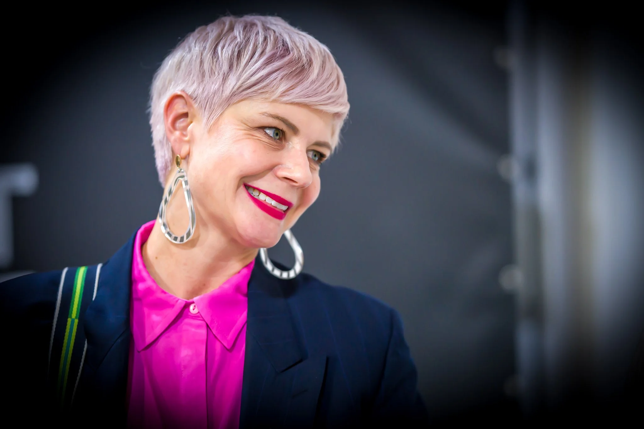 Une personne souriante avec une coiffure courte et blonde, portant des boucles d'oreilles larges et un vêtement rose et bleu.