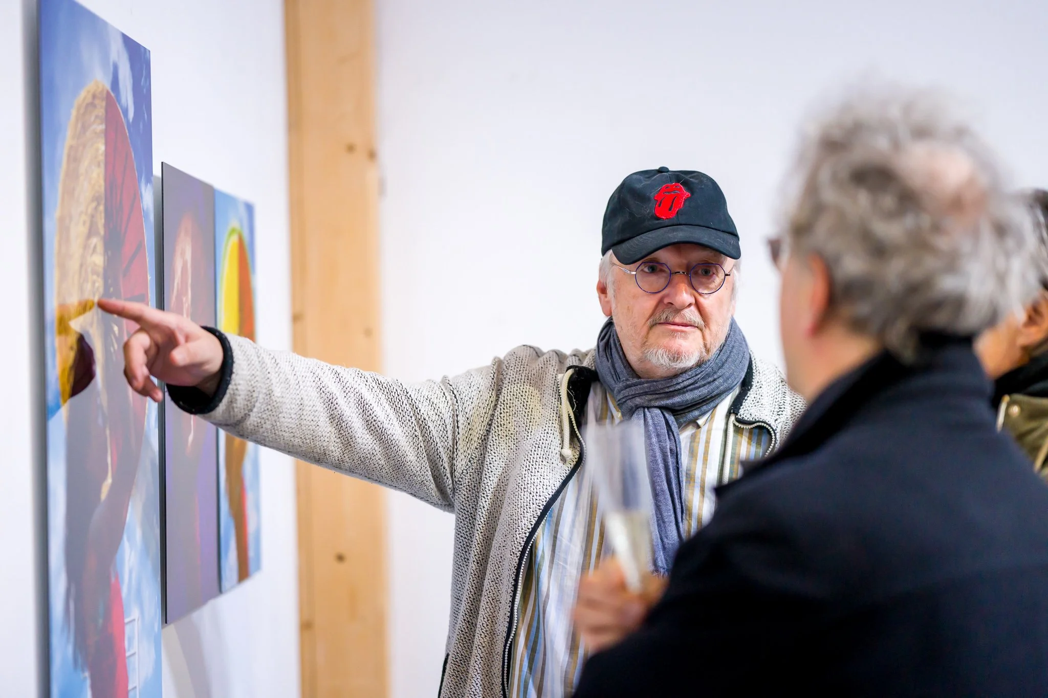 Un homme portant une casquette noire et des lunettes montre du doigt une peinture accrochée au mur, discutant avec un autre homme. Il porte une écharpe et un manteau. Des œuvres d'art colorées sont visibles en arrière-plan.