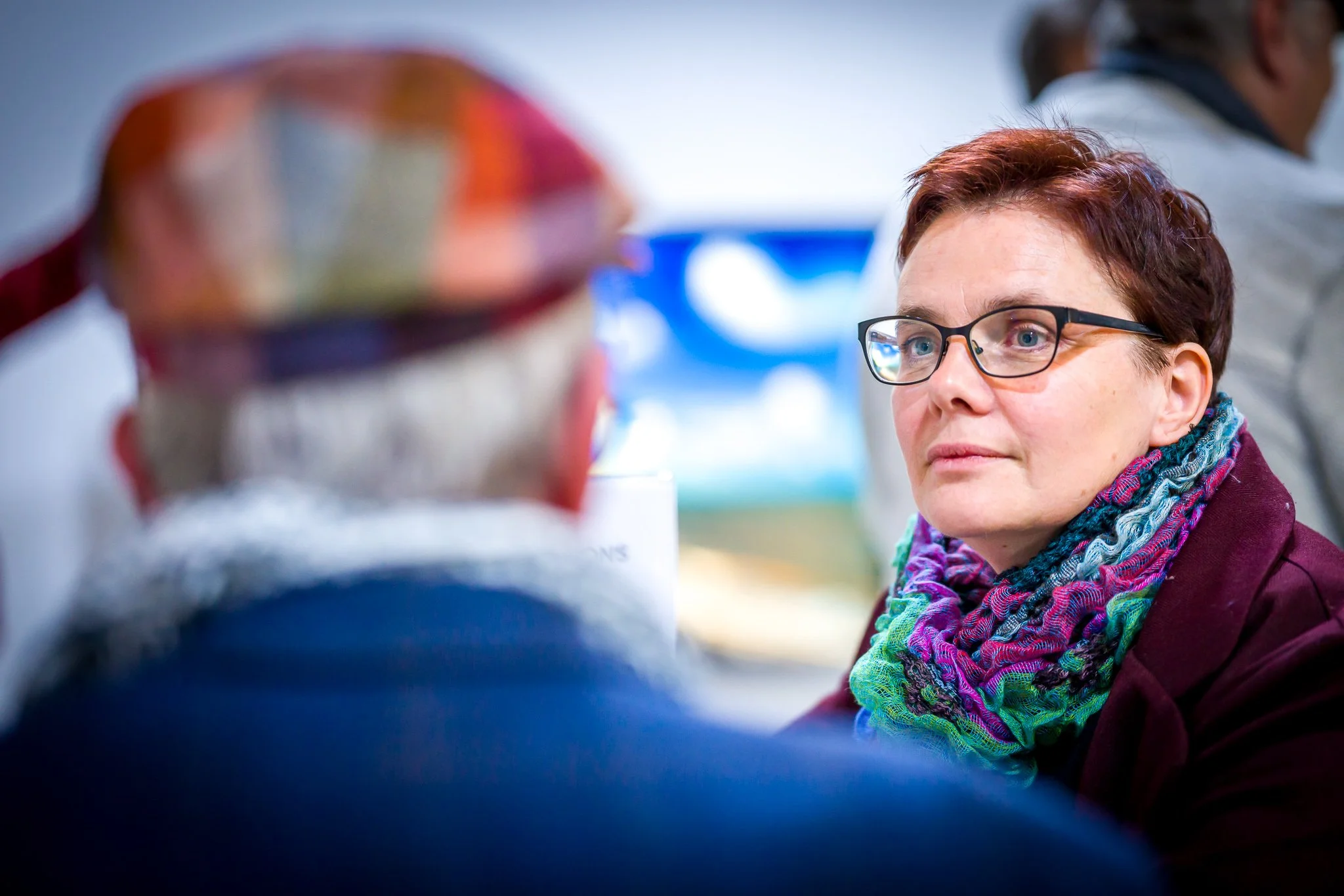 Une femme portant des lunettes et une écharpe colorée regarde une autre personne au premier plan floue.