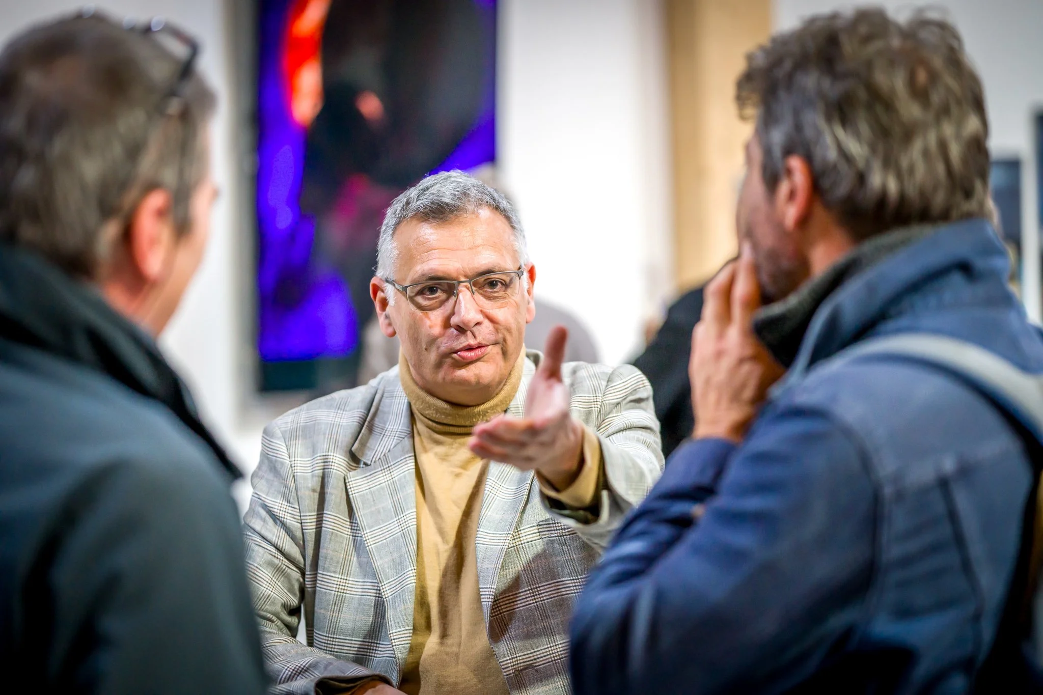 Trois hommes en discussion, l'un d'eux parlant avec un geste expressif de la main, avec un fond d'exposition artistique flou.